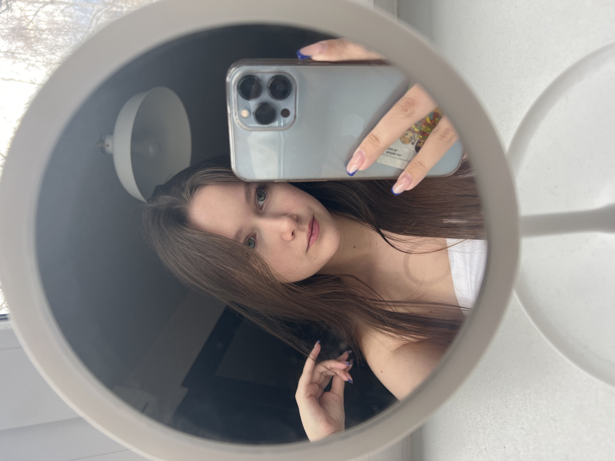 This media file captures a mirror selfie of a young woman, likely taken in Chaikovsky, Russia, during the daytime. The primary subject is a woman with long, straight brown hair, light eyes, and pinkish lips, looking directly into the mirror. She is wearing a white top or bra strap.
The image is framed by a round, light-colored mirror. The woman is holding a light blue or clear-cased iPhone (identifiable by its triple camera array, likely an iPhone 13 Pro or 14 Pro) to take the picture. Her nails are long, styled with a French manicure and distinct blue tips. On the back of her phone case, a small sticker featuring cartoon-like figures is visible, though any text on it is too small to discern. Her other hand is posed near her hair.
In the reflection, to the left of her head, a white, bell-shaped light fixture or lamp shade can be seen. Beyond the mirror's frame, a blurred view of bare tree branches against a bright sky suggests an outdoor scene through a window, indicating the photo was taken indoors. To the right, another white, curved object, possibly part of a second mirror or a stand, is visible. The overall lighting is bright, consistent with natural light from a window during the day, possibly winter or early spring given the bare trees. The woman's expression is calm and neutral.