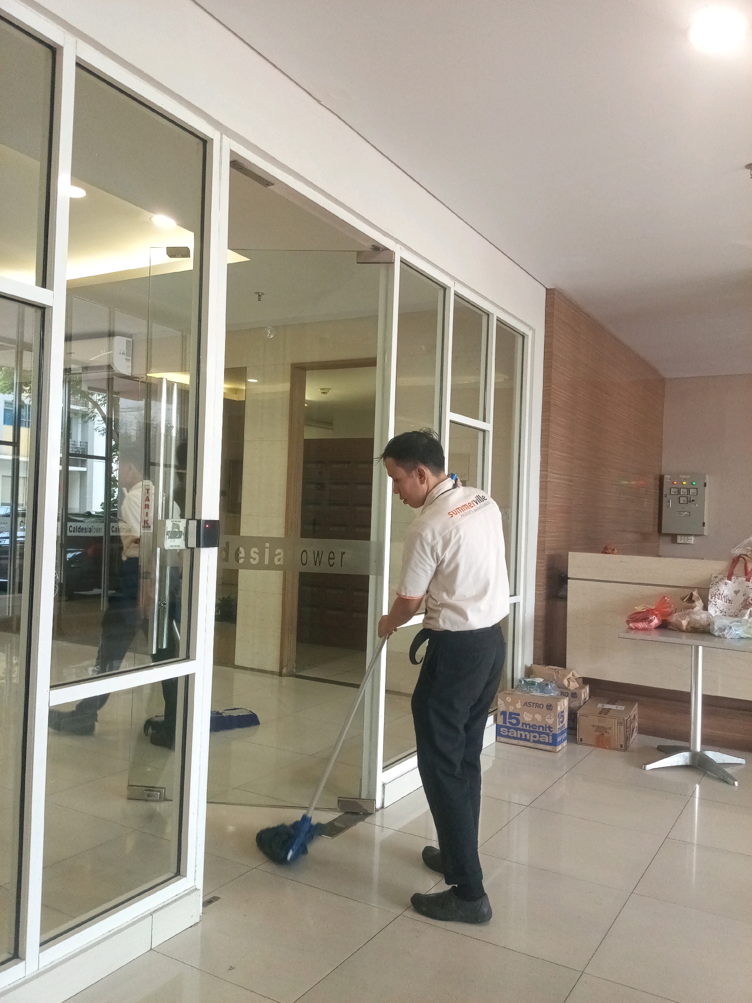 The image depicts a man in a light-colored uniform with the logo "summerville" on the sleeve, mopping a tiled floor in what appears to be the entrance or lobby of a building. The man is wearing black pants and dark shoes. He is holding a blue mop with a metal handle. The building has large glass doors and windows, with reflections of the interior visible. There are signs on the glass doors, one reading "Caldelsia tower". In the background, there is a table with some bags and boxes on it, and an electrical panel mounted on a wall with wood-paneled accents. The scene is indoors, with artificial lighting. The general environment suggests a commercial or residential building in an urban setting, possibly in Bekasi, Indonesia, given the mention of "Caldelsia tower" which may be a local landmark.