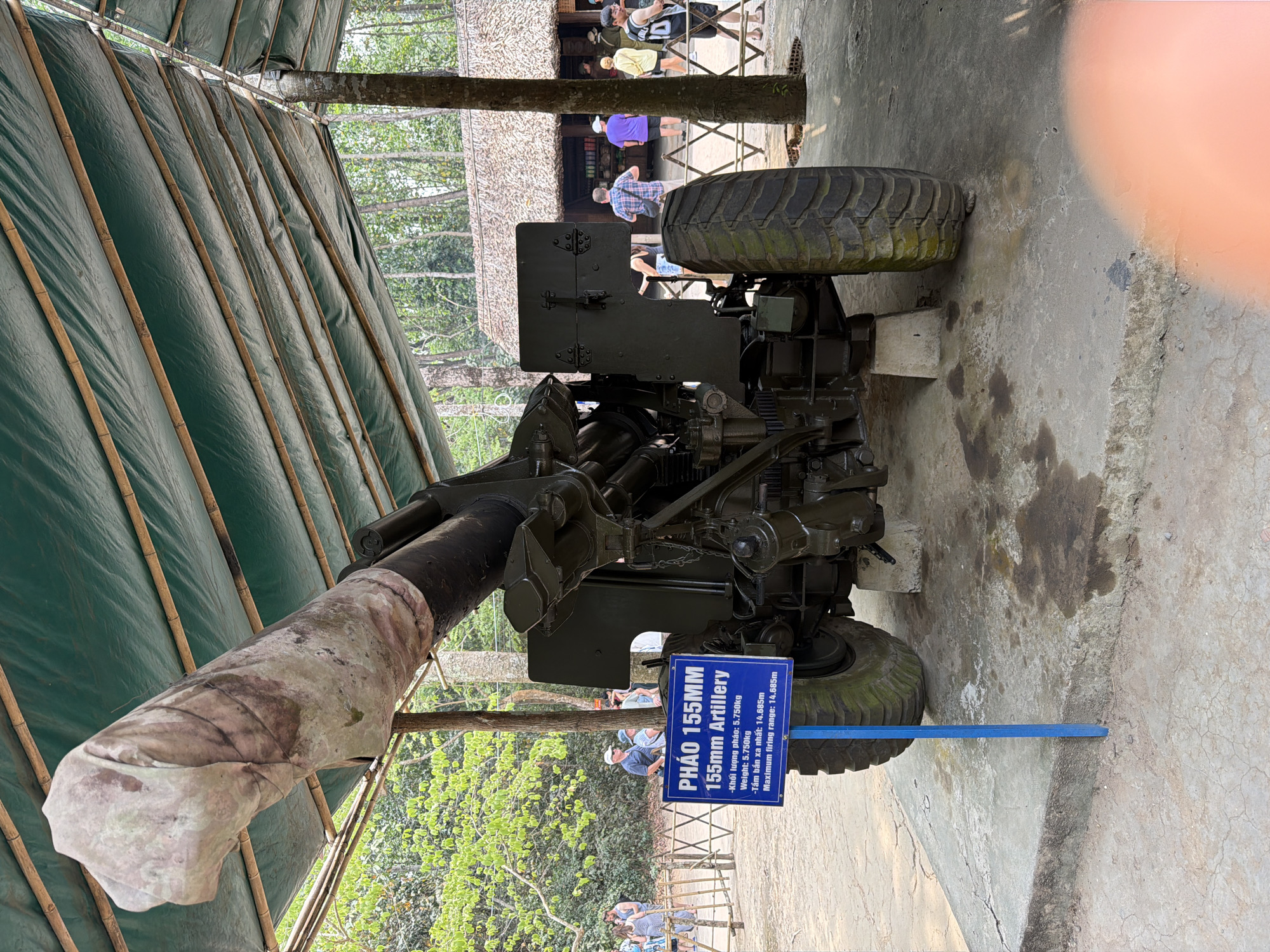 A daytime image captures a large, dark green 155mm artillery piece displayed outdoors, likely at a historical site or museum in Ho Chi Minh City, Vietnam. The artillery rests on a concrete surface, partially sheltered by a green tarpaulin canopy supported by bamboo poles. Its long barrel is encased in a light-colored, possibly camouflaged, fabric cover. The weapon features robust, heavily treaded tires.
A blue informational sign stands beside the artillery, providing details in both Vietnamese and English. The text reads: "PHÁO 155MM / 155mm Artillery," and lists its specifications: "Weight: 5.750kg" (Khối lượng pháo: 5.750kg) and "Maximum firing range: 14.685m" (Tầm bắn xa nhất: 14.685m).
In the background, several casually dressed people, likely visitors or tourists, are seen observing the exhibits or moving through the area. Behind them, verdant trees and dense foliage are visible, along with traditional thatched-roof structures, suggesting a natural and culturally relevant setting. The clear daylight indicates pleasant weather, and the scene depicts an educational or historical tour activity.