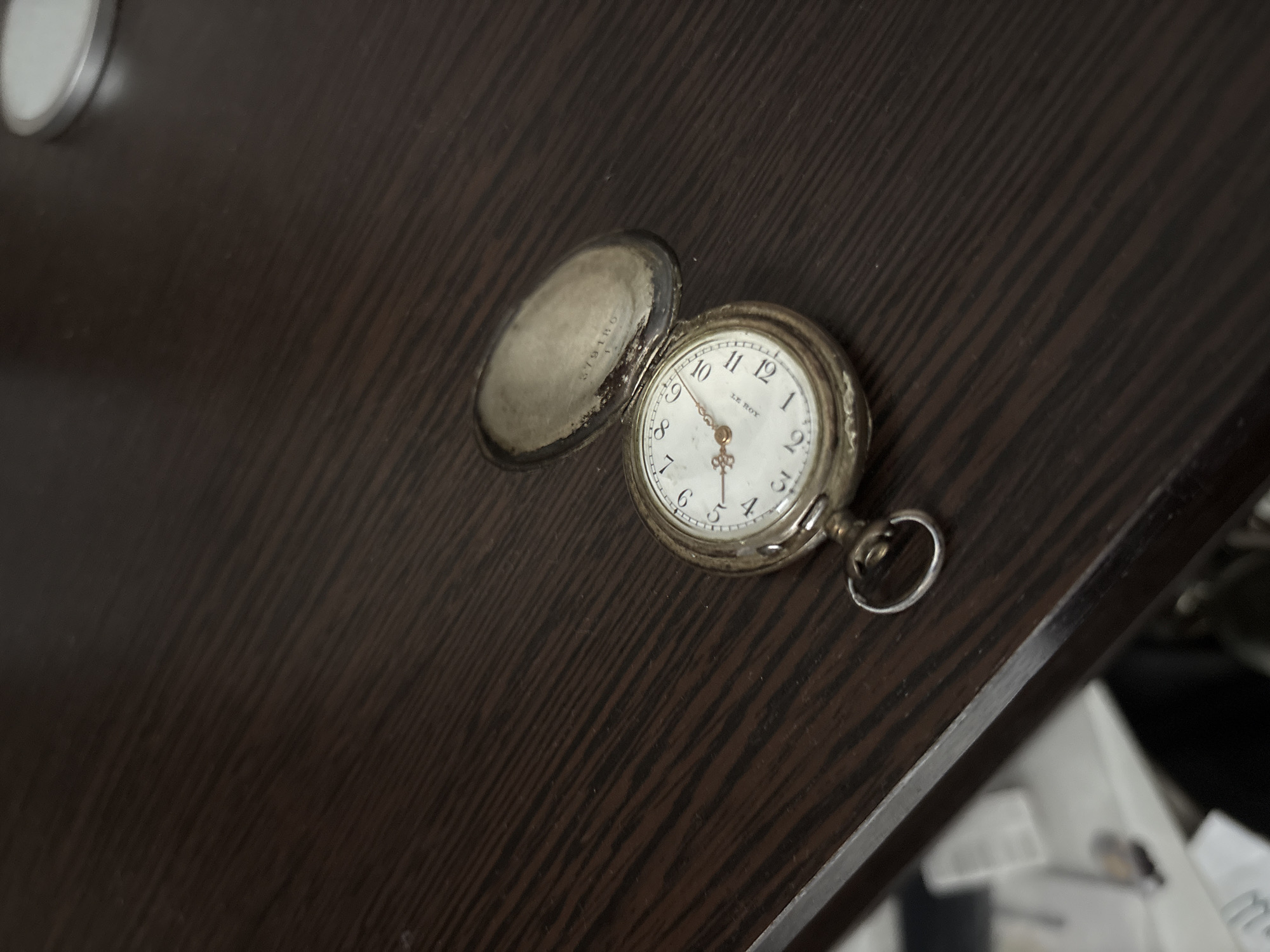 A vintage pocket watch rests open on a dark wood surface. The watch face is white with black Arabic numerals and two golden hands indicating the time. The watch cover is open, revealing an inscription inside. The background is a textured, dark brown wooden surface with diagonal lines. The image is taken from a slightly elevated angle, with the pocket watch positioned in the lower left quadrant. There is a suggestion of a blurred object to the left of the watch, possibly a book or papers, and a white circular object with a silver rim in the upper right corner. The scene appears to be indoors.
