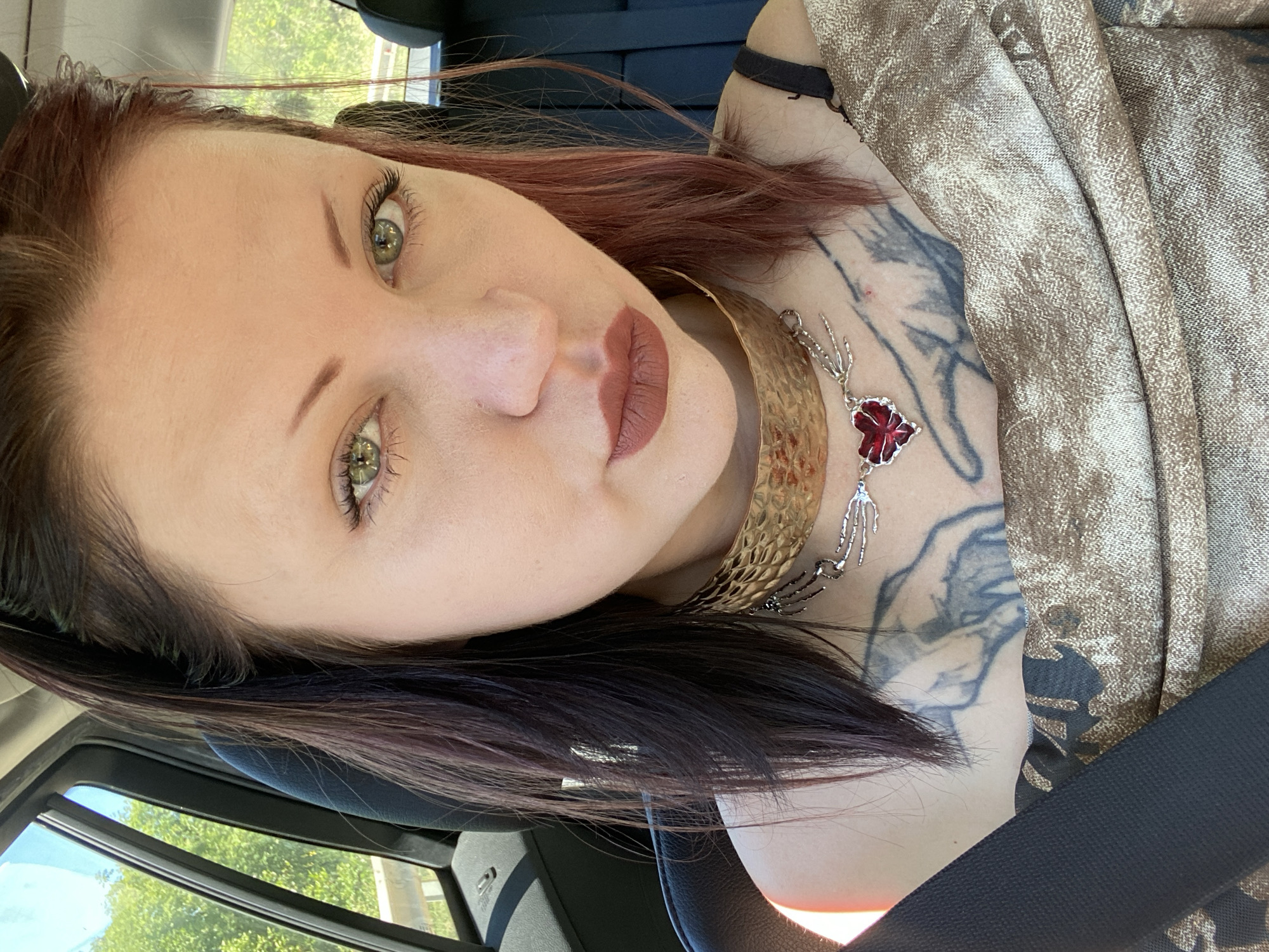 A woman is inside a car wearing a seatbelt. She has reddish-brown hair, green eyes, and is wearing lipstick and a gold-colored necklace. A tattoo is visible on her chest. The location in the city of Kruče - Kërruçi, Montenegro cannot be determined from the image.