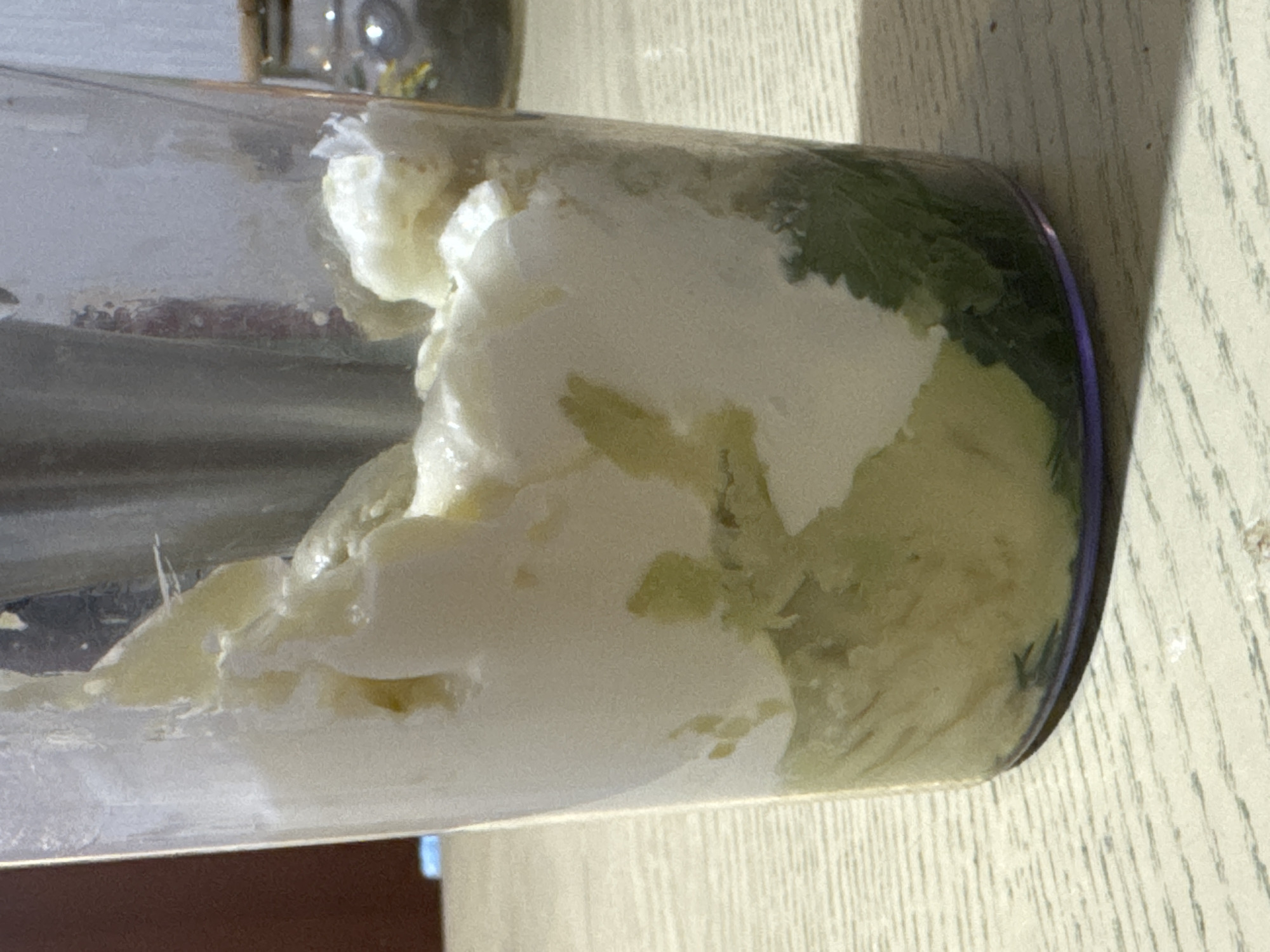 A glass containing a mixture of white and green substances rests on a light brown wooden surface. The white substance appears creamy and occupies the forefront, while the green substance is visible toward the back and right side of the glass. The glass is partially transparent, allowing a glimpse of the substances within. A blurred object is visible in the background above the glass. The wooden surface has a subtle grain pattern and is illuminated by bright sunlight. No people are visible. The image does not provide sufficient information to determine the location as Chaikovsky, Russia.