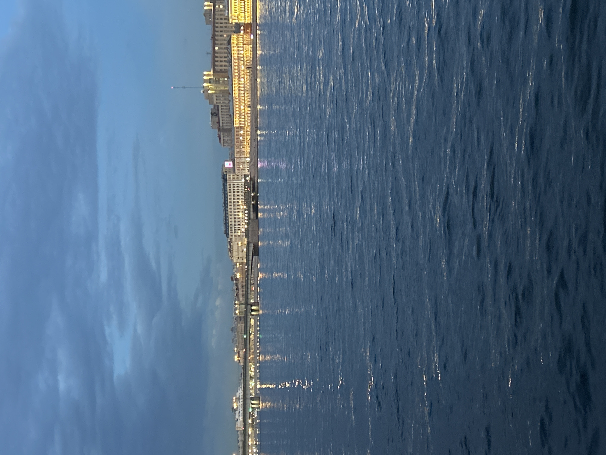 The image shows a wide river with buildings and a bridge in the background. The sky is dark blue with some clouds. The buildings are lit up with yellow lights, and their reflections can be seen in the water. The river is dark blue and appears to be slightly choppy. The scene suggests it is dusk or early evening.