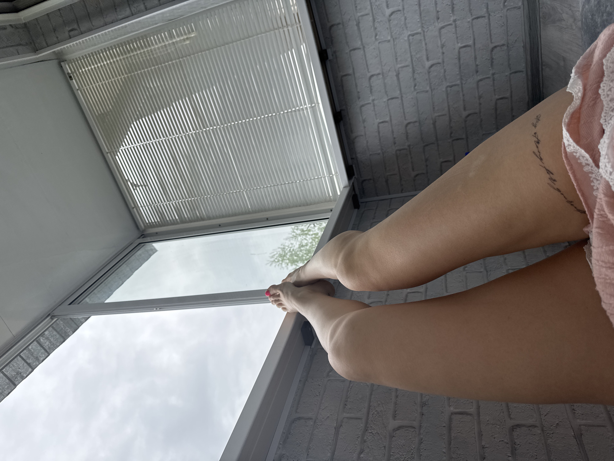 The image shows a view from inside a room, looking out through a window. The person's legs are visible in the foreground, with a tattoo on the thigh. A pink garment is partly visible. The window is open, offering a view of a cloudy sky. The wall and ceiling are visible, featuring a faux brick pattern and white blinds respectively. The specific location within the city of Chaikovsky, Russia, cannot be determined from this image.