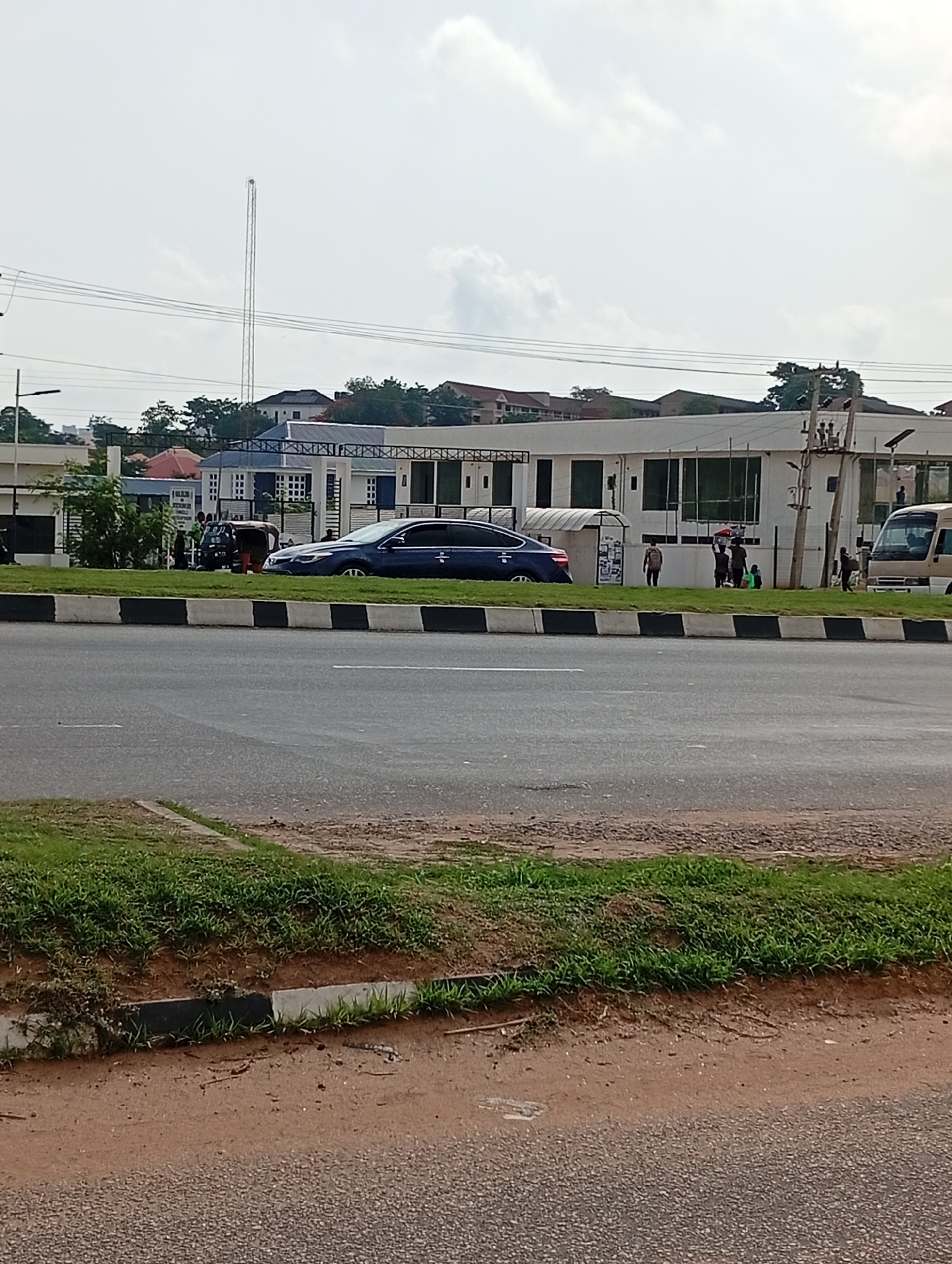 The image depicts a street scene in Aso, Nigeria. A dark blue sedan is parked on the side of the road, next to a three-wheeled vehicle with a small group of people nearby. Further down the road, there is a group of people standing near a white van. In the background, there is a building with a white facade and large windows. Behind the building, there are trees and rooftops of other houses. A tall communication tower is visible in the distance. The road is divided by a black and white curb. The foreground shows a grassy area with a dirt path. The sky is overcast.