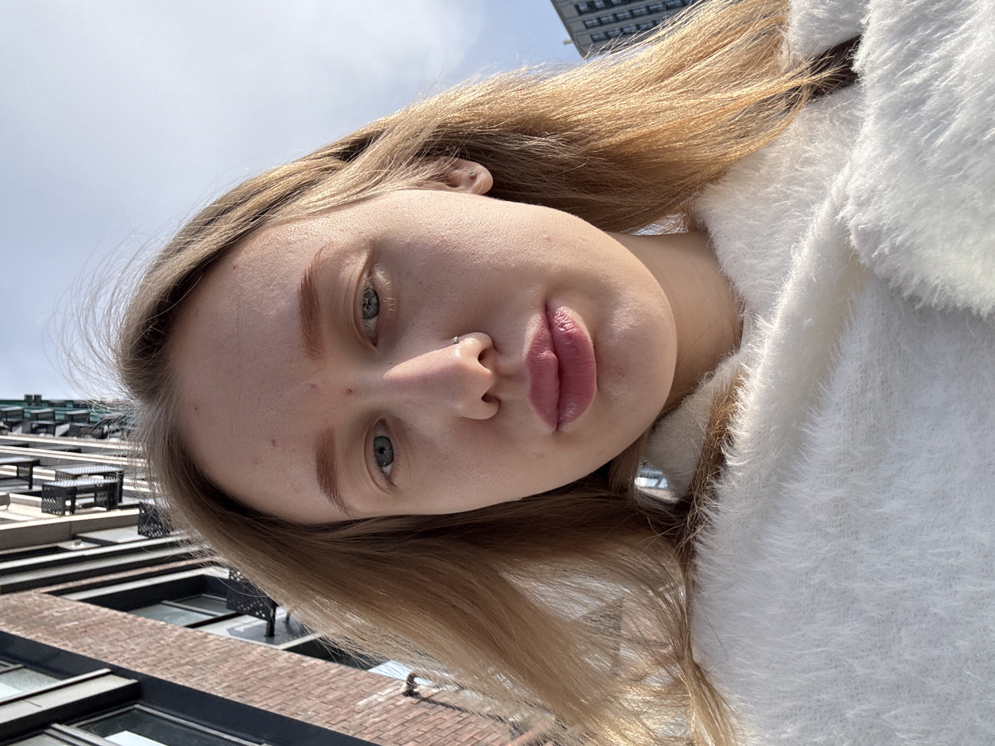 A young fair-skinned woman is facing the camera with her head tilted to the right. She has blonde hair and her complexion is clear save for a few blemishes on her forehead and nose. She has light eyes and pink lips. She wears a white fluffy garment. The background includes parts of buildings, including a multi-story brick structure with windows and what appear to be small tables or platforms on the rooftop. Part of a taller building can be seen in the distance. The location is ambiguous with no indication that the location is in the city of Ovoshchnoy, Russia.