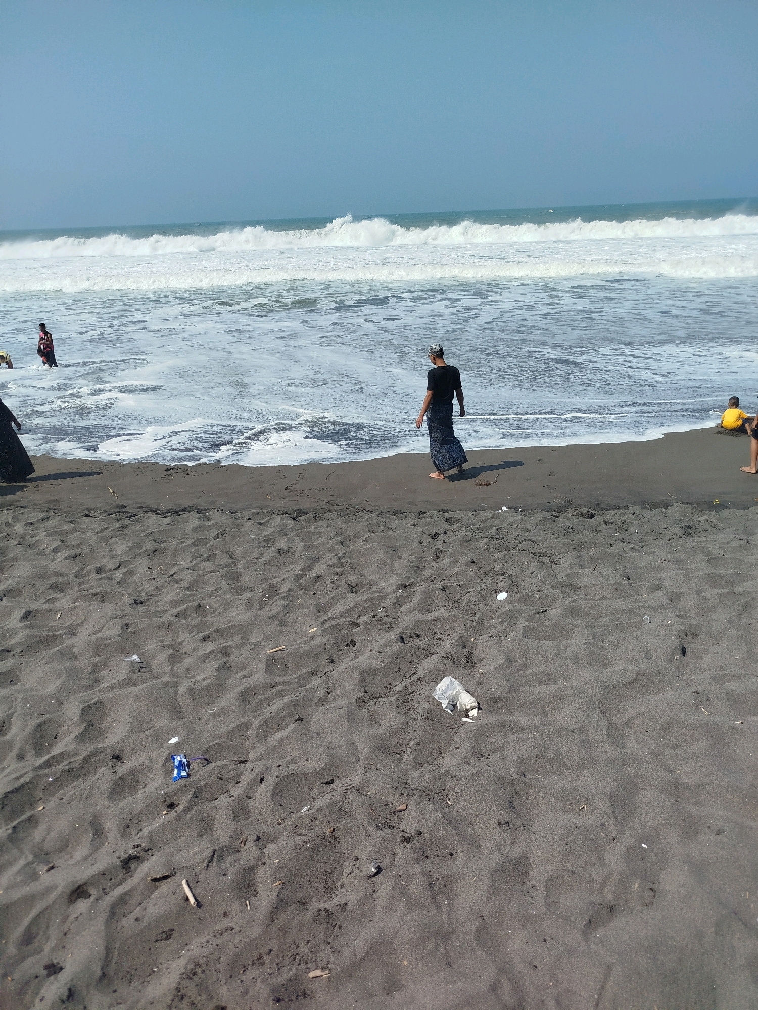 The image depicts a beach scene at Kenoyajaya, Indonesia. The foreground is dominated by dark, fine-grained sand with scattered debris, including a white plastic bag and a small blue and white object. In the middle ground, the dark sand meets the ocean with waves breaking. Several people are visible near the water. One person, wearing a black t-shirt and a patterned sarong, walks along the shoreline. To the left, a person in a red and black garment stands in the shallow water, and another person in a yellow shirt is seated on the sand. Further out, waves crash, creating white foam. The sky is clear and pale blue.
