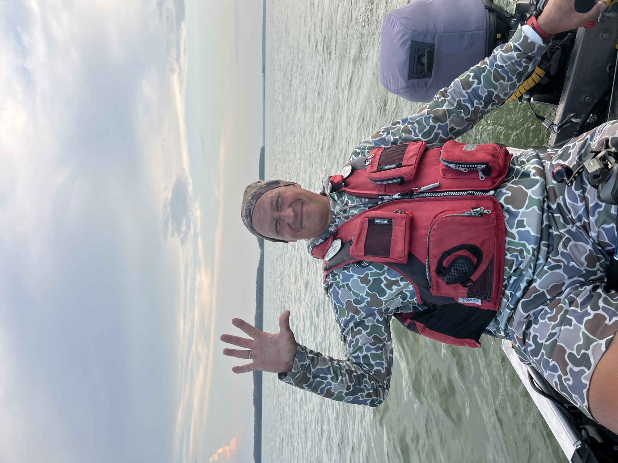 An adult male wearing a camouflage long-sleeved shirt, a red life vest, and a camouflage beanie is sitting in a boat on a body of water. He is smiling and holding up his right hand with his palm facing the camera, displaying five fingers. The boat has an outboard motor at the rear, partially covered by a gray tarp. The water is choppy, and a tree line is visible in the distance across the water. The sky is overcast with scattered clouds. The location is not specified as Novopichugovo, Russia.