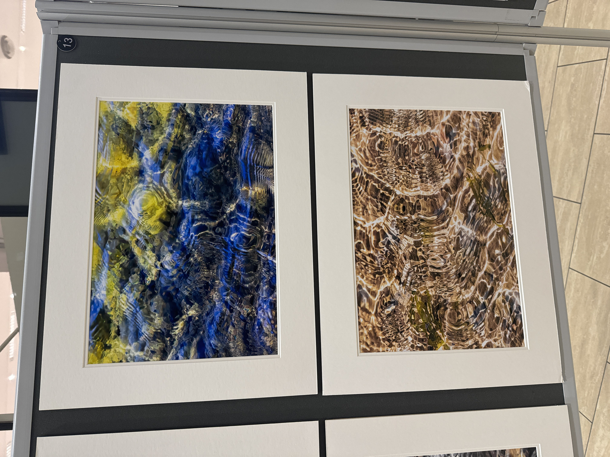This is a photograph of an art exhibition, likely within the city of Kilkenny, Ireland. The image displays two framed photographs mounted on a dark grey partition. Both photographs feature abstract close-up shots of rippling water, with the top image predominantly showcasing blues and yellows, and the bottom image featuring browns and earthy tones. A small circular tag with the number "13" is visible on the top right of the partition, indicating the artwork's number. The lighting suggests an indoor setting, possibly a gallery or exhibition space, with the floor tiles visible in the foreground. There are no people or specific landmarks of Kilkenny visible.
