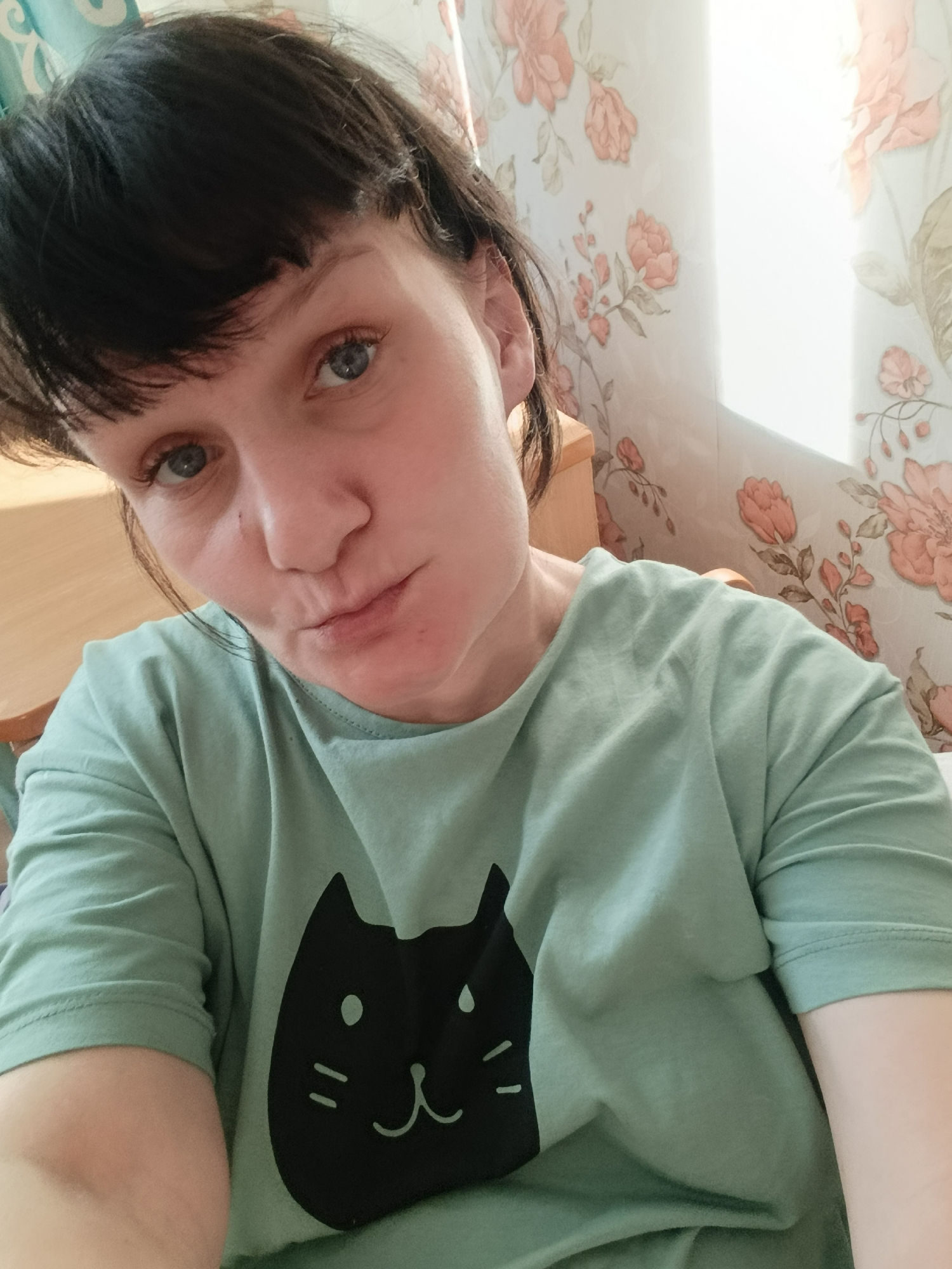 A woman with short dark hair and blue eyes is taking a selfie. She is wearing a light green t-shirt with a black cat graphic. The background consists of a wooden surface and floral patterned curtains. There is no evidence to confirm the location in the city of Фоки, Russia.
