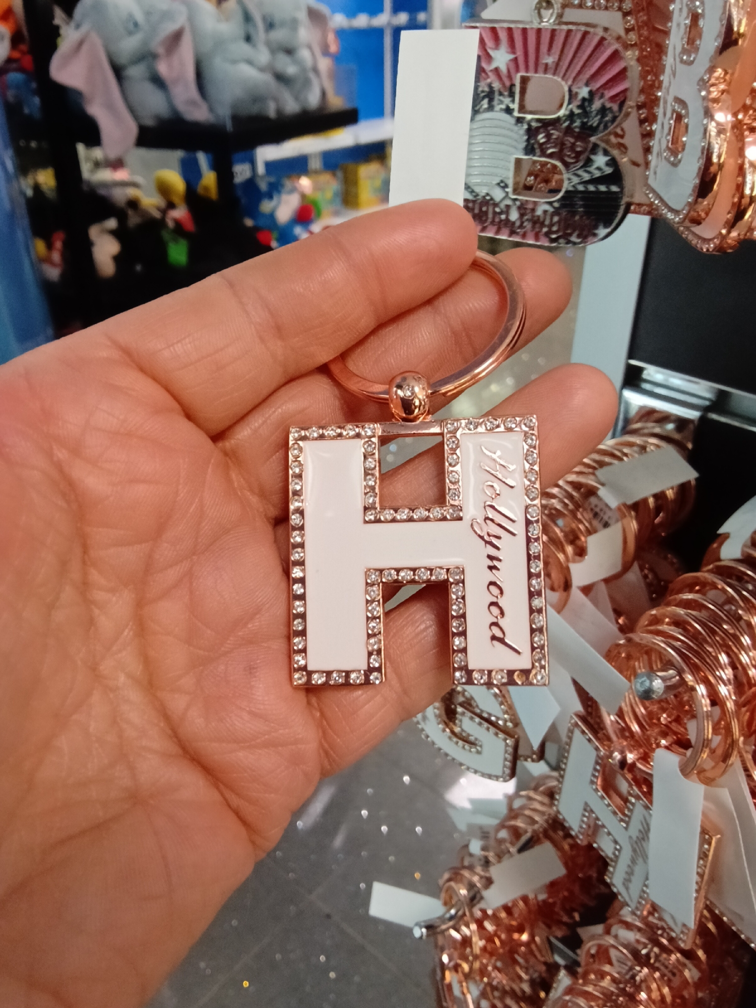 A human hand, appearing to be an adult's, holds a rose gold-toned metal keychain in the foreground. The keychain is shaped like the capital letter "H," featuring a white enamel inlay bordered by small, clear, faceted stones. The word "Hollywood" is inscribed vertically in a cursive script on the right vertical segment of the "H." A circular rose gold-toned keyring is attached to the top of the letter.
To the right, a retail display rack is visible, holding numerous similar rose gold-toned keychains, some displaying letters such as "G" and "H" with "Hollywood" branding. These items are attached to individual white tags and suspended from metal hooks.
The background to the left is out of focus, showing multiple shelves stocked with merchandise. The uppermost shelf contains several light-colored plush toys, possibly elephants, with light pink inner ear accents. Other colorful, indistinct items are visible on lower shelves, consistent with a retail store environment.
Based on the explicit "Hollywood" branding on the keychains, the scene is located within a souvenir or gift shop in the Hollywood district of Los Angeles, California, United States of America.