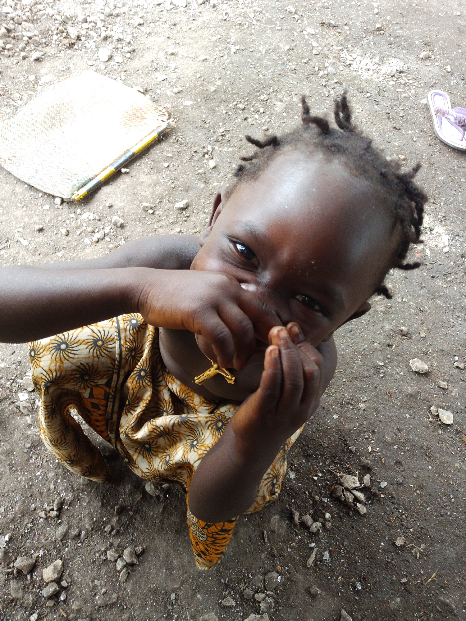A young child with dark skin and short, dark, curly hair, is shown from a high-angle shot. The child is wearing a patterned garment in shades of yellow and orange with a black design. The child's hands are raised to their face, obscuring their mouth and nose. A small, gold-colored crucifix necklace is visible around the child's neck. The child is positioned on the ground, which appears to be dirt or gravel mixed with small stones. In the background, a woven mat, possibly a fan, lies on the ground to the left, and a single purple sandal is visible to the right. The overall environment suggests an outdoor setting in a possibly rural or less developed area. The location is identified as Otulu, Nigeria.
