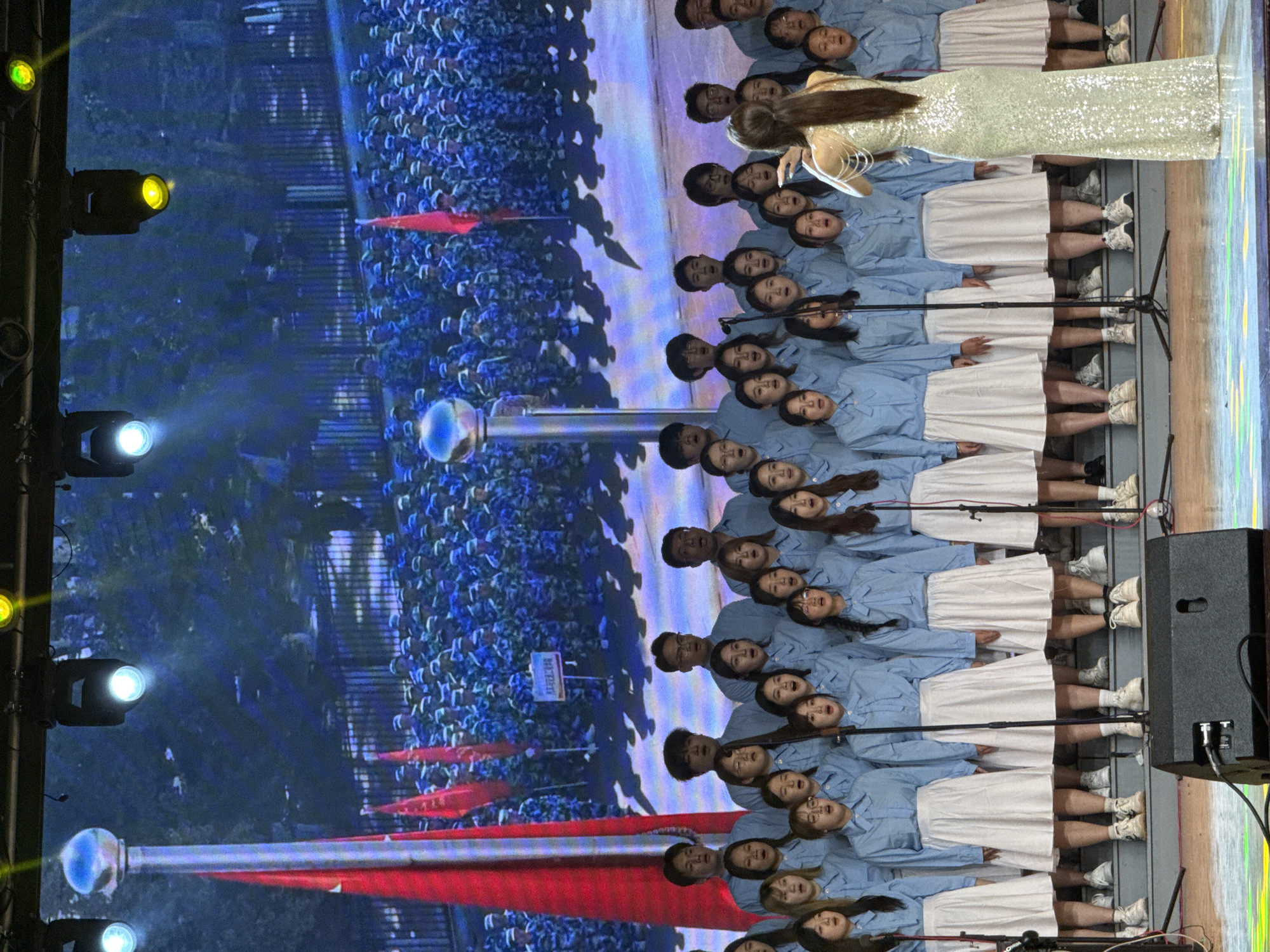 The image captures a vibrant performance on a stage, likely indoors, with a massive LED screen forming the backdrop. In the foreground, a choir, predominantly composed of young individuals, stands in multiple rows. They are uniformly dressed in light blue long-sleeved shirts, white skirts, and white sneakers, with many mouths open in song. A female soloist, with long dark hair and wearing a sparkling, light-colored long gown, stands slightly to the left of the choir, holding a microphone and performing.
On the left side of the stage, several spotlights emit yellow, green, and white light. A large black speaker box is visible on the right. The LED screen behind the performers displays a scene of a vast historical or commemorative gathering, featuring numerous individuals, many holding red flags, and what appears to be a large flagpole. A prominent white sign with dark Chinese characters "抗美援朝" (Kàng Měi Yuán Cháo), which translates to "Resist US Aggression and Aid Korea," provides a strong thematic context related to the Korean War.
The setting, enhanced by the stage lighting, suggests an evening event or a performance in a darkened venue. The performers exhibit focus and engagement in their singing. This event, taking place in Guta, China, appears to be a patriotic or commemorative concert.