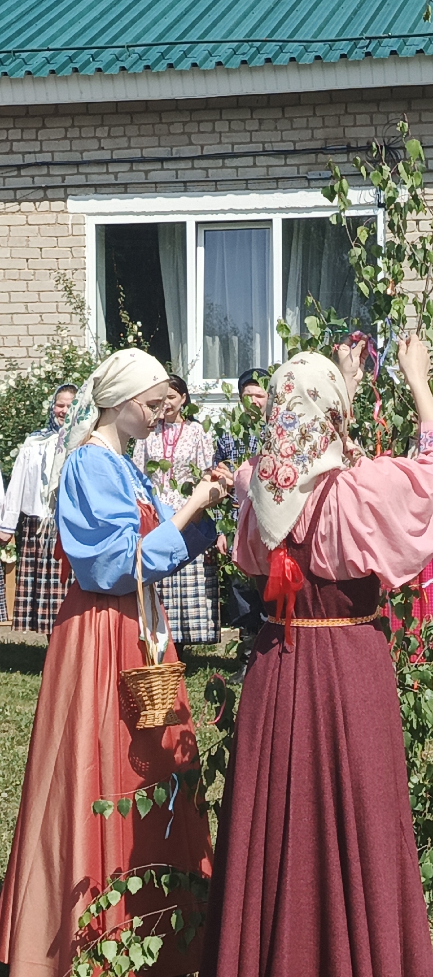 Two women in traditional Russian clothing are decorating a tree with colorful ribbons. They are standing in front of a brick building with a green roof and a white window frame. Other people, also in traditional attire, stand behind them, seemingly observing the ritual. One woman carries a small wicker basket. The scene appears to be taking place outdoors in a grassy area. Based on the location data, this is likely in the city of Кемуль, Russia.