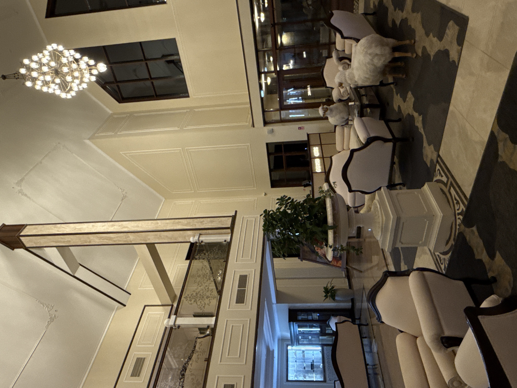 The image depicts the interior of a grand lobby, possibly a hotel or a similar establishment, with high ceilings and ornate architectural features. A large, multi-tiered chandelier hangs from the ceiling, illuminating the space. The walls are a pale yellow with decorative molding. In the foreground, several plush, cream-colored armchairs and sofas are arranged around a dark coffee table. Two sheep figures, appearing to be decorative statues or mascots, are placed near the seating area. One sheep stands facing the camera, while the other is partially obscured. A large potted plant sits on a decorative pedestal to the left of the sheep. The flooring appears to be a combination of marble tiles and a dark, patterned rug. In the background, a reception desk is visible, with glass doors leading further into the building. A second-floor balcony with decorative railing overlooks the lobby. The lighting suggests it is either evening or the interior lights are the primary source of illumination. The environment is clean and well-maintained, with a formal and luxurious aesthetic. There are no visible people in the scene. There is no information available about the city of Urlu-Aspak, Russia.
