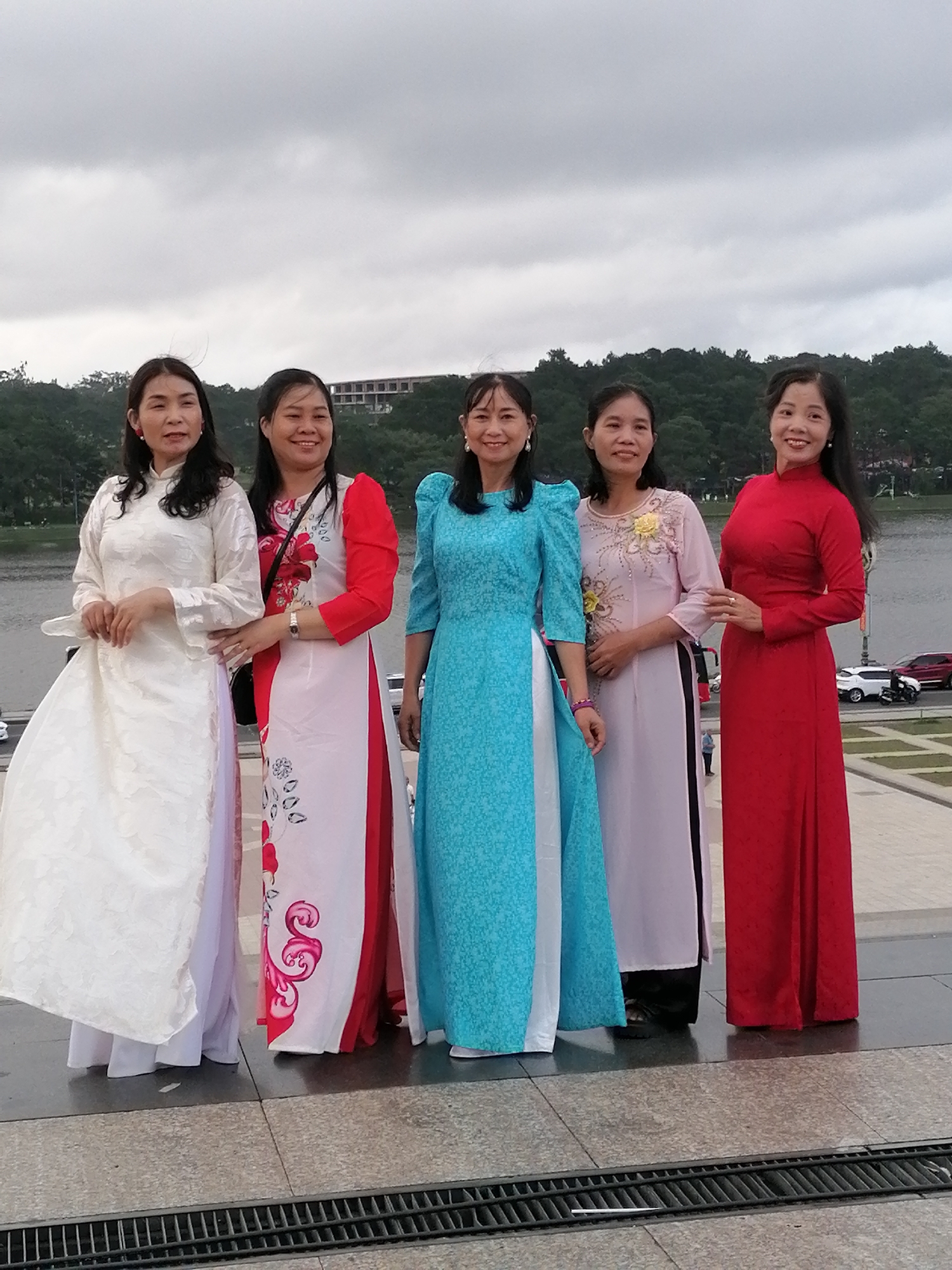 Five Vietnamese women are posing for a photograph in front of a body of water with a wooded hillside and buildings in the background. The women are all wearing traditional Ao Dai dresses in various colors: white, red and white, blue, pink, and red. They are standing on a paved area with a metal grate in the foreground. The location is likely the Xuan Huong Lake in Da Lat, Vietnam, a popular tourist destination known for its scenic beauty and colonial architecture. The overcast sky suggests a cloudy or rainy day.