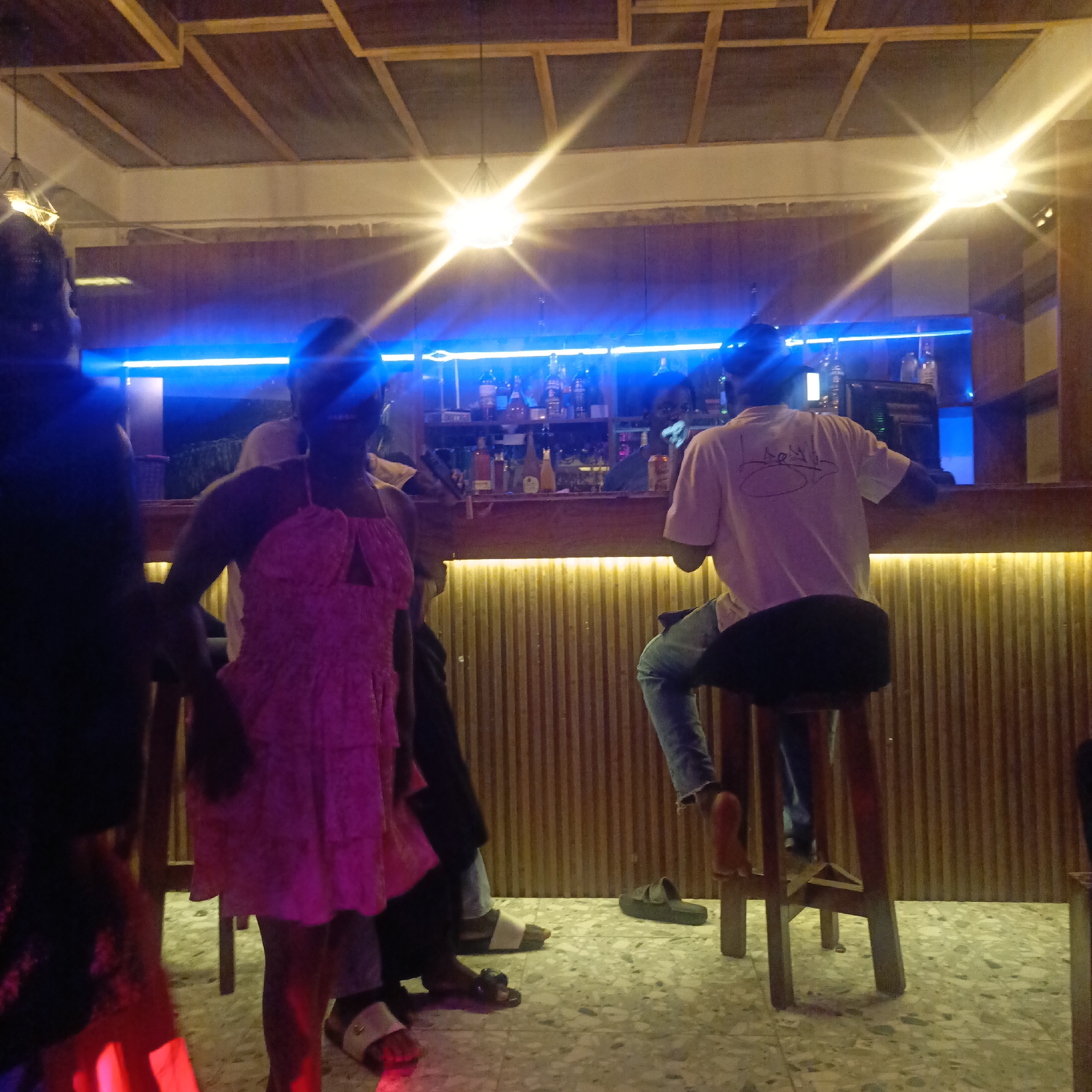 Here's a detailed description of the image:
**Setting:** Indoor bar setting in Akwanga, Nigeria. The bar features a counter with vertical wood paneling, a blue neon light strip above, and shelves stocked with bottles of alcohol. The ceiling has a wooden grid design and hanging lights. The floor has a speckled tile pattern.
**People:** Several people are present. A woman in a pink patterned dress stands near the bar. A man wearing a white t-shirt and jeans is sitting on a wooden stool at the bar. Other individuals are seen standing or sitting near the bar but are not distinctly visible.
**Objects:** Visible objects include bar stools, bottles of alcohol on the shelves behind the bar, and flip-flops/sandals worn by individuals.
**Actions/Interactions:** The man at the bar is sitting. The woman is standing. Other people are partially seen and potentially engaging in conversations or observing the scene.
**Environment:** The lighting is a mix of bright white light, blue neon light, and some red ambient light. The overall atmosphere appears lively and social.