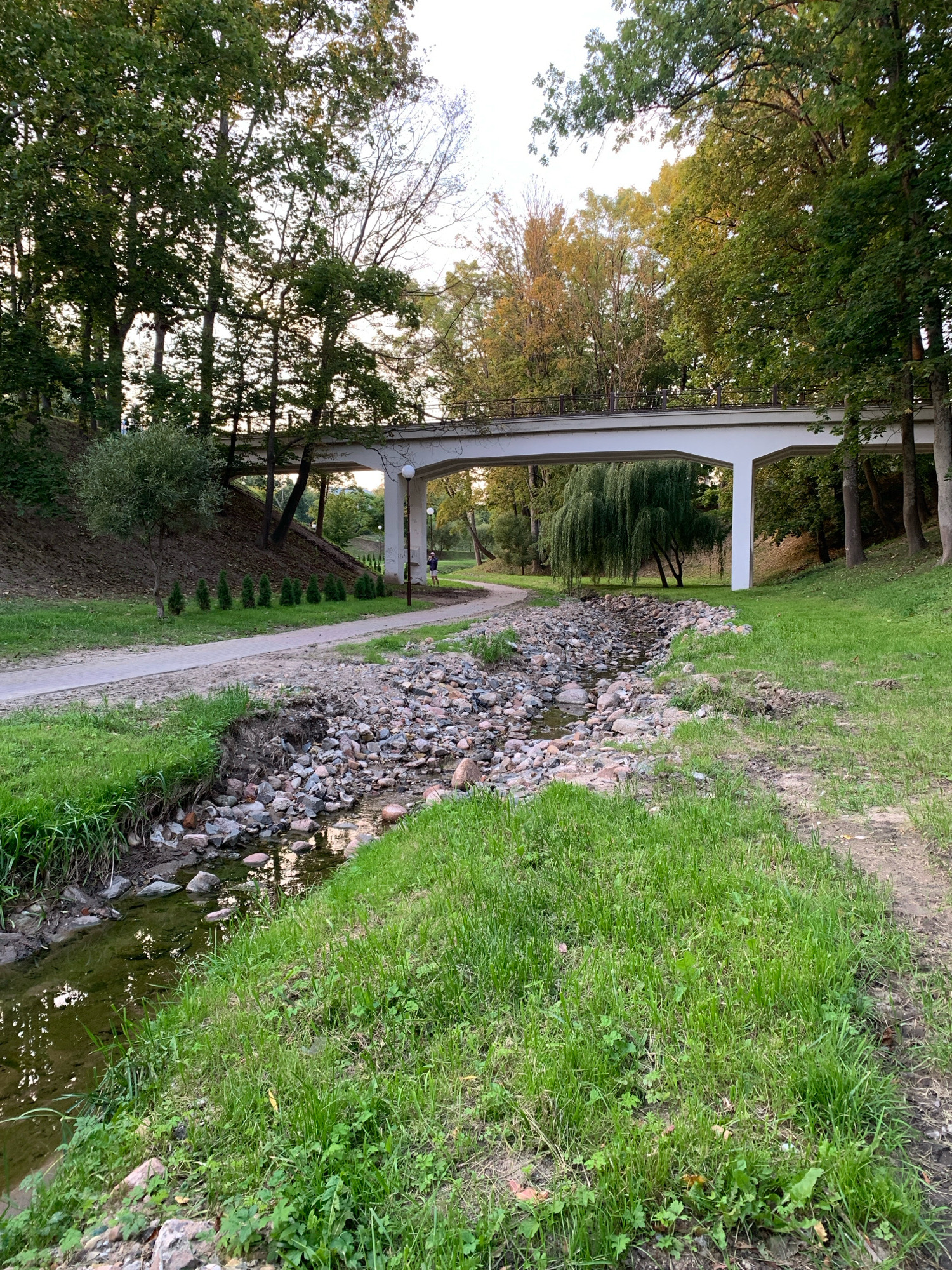 A serene park scene in the city of Hrodna, Belarus, captures a dry creek bed lined with stones, flanked by lush green grass. A paved walking path curves gently to the left, leading towards a modern concrete bridge that spans the scene. Tall trees with full green foliage and some with autumn colors frame the background. A solitary figure can be seen walking on the path under the bridge. The overall atmosphere is peaceful and natural.