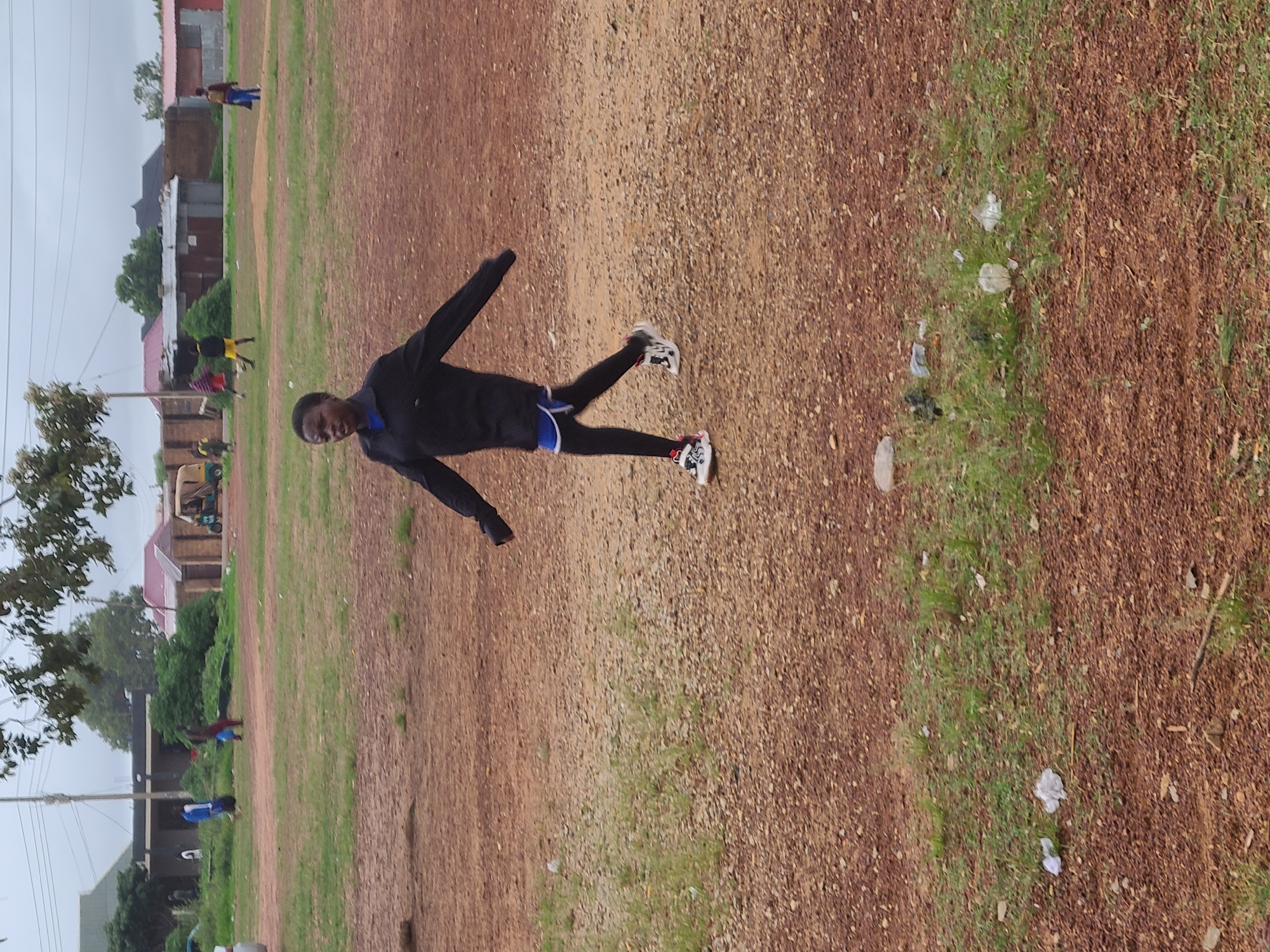 The image captures an outdoor scene in Rafin Bauna, Nigeria, under an overcast sky. The primary subject is a young man, appearing to be in his late teens or early twenties, seen mid-stride, suggesting he is running or jogging. He is positioned in the center-left, facing slightly towards the viewer and to the right. He wears a black hooded jacket, black athletic leggings with blue shorts visible underneath, and multi-colored athletic shoes with white, black, and red accents. His expression is neutral, with a hint of a smile.
The setting is a large, open field of reddish-brown earth, resembling a dirt track or unpaved ground. A strip of sparse green grass runs through the field, notably visible on the right side and in the middle ground. Scattered light-colored trash, such as plastic bags and wrappers, is visible on the ground, especially within the grassy patches.
In the background, on the left side, several other individuals are visible, some walking or standing near structures. Simple, low-rise buildings and fences, some with corrugated metal roofs, define the distant skyline, indicating a semi-urban or rural environment. A few trees and power lines are also present. A light-colored three-wheeled vehicle is partially visible in the far left background. The time of day appears to be daytime, possibly during a cloudy period. No visible text is present in the image.
