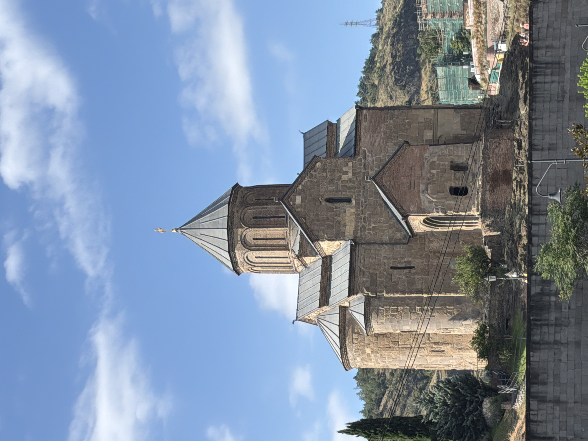 The image captures the Sameba Cathedral in Tbilisi, Georgia, on a clear day with a bright blue sky and scattered white clouds. The magnificent stone cathedral is the central focus, with its distinctive conical dome topped by a golden cross. The structure is made of large, roughly hewn stones, and features multiple rounded towers and arched windows. A smaller, brick-built annex is visible to the right of the main structure.
The cathedral is situated on a hillside, with a steep, tree-covered slope rising behind it. In the distance, a few modern buildings and construction sites, partially covered in green netting, are visible, suggesting ongoing development or renovation in the area. To the left of the cathedral, a tall, slender cypress tree stands out against the landscape.
In the foreground, a stone retaining wall runs along the bottom of the image, with some greenery and trees growing over it. A streetlamp is positioned on the wall, and a few indistinct figures of people can be seen in the middle ground on a pathway or terrace, observing the cathedral or the surroundings. Electrical wires are strung across the scene, partially obscuring the cathedral and the hillside. The overall impression is one of a historical and religious landmark set against a backdrop of both natural and urban elements.
