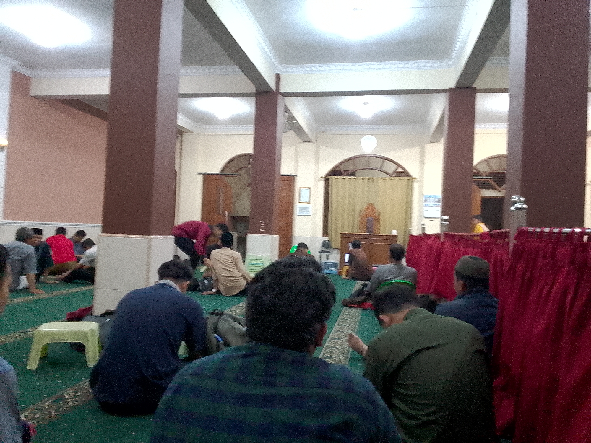 This image depicts the interior of a mosque in Kertajaya, Indonesia. Several individuals are present, engaged in various activities. In the foreground, a person wearing a dark blue shirt is seated facing away from the camera, with another person in a green shirt to their right. To the left, a light yellow stool is visible on the green carpet.
In the mid-ground, a man in a dark blue shirt is seated, facing towards the right. Further back, a group of men are seated on the carpet. Some are facing towards the front of the mosque, where a red curtain is partially visible. Other individuals are either standing or in the process of sitting down. The mosque features large, dark brown pillars and a white ceiling with illuminated panels. Arched doorways and wooden furniture are also present in the background. The overall scene suggests a gathering or prayer session within the mosque.
