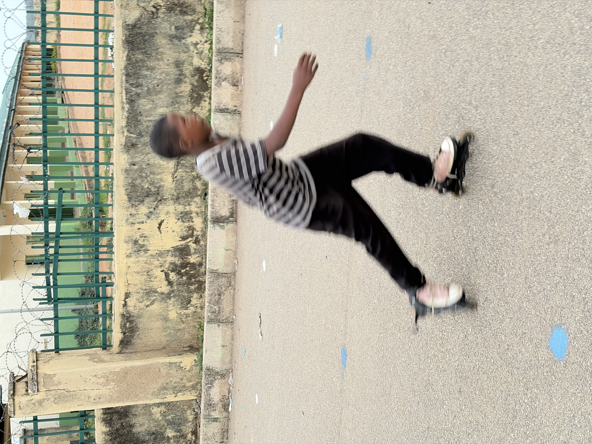 A person is inline skating on an asphalt surface. The person is wearing a striped shirt, black pants, and inline skates. The asphalt surface has some white and blue markings on it. To the left of the skater is a building with a green fence. The building and fence appear to be old and slightly dilapidated. The location is presumed to be in the city of Jos, Nigeria. Due to motion blur, the person's actions are indistinct.