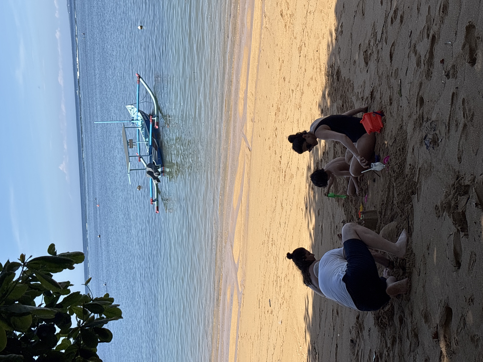 The image shows a beach scene in Denpasar, Indonesia. On the left, a calm ocean with a traditional Indonesian outrigger boat anchored near the shore. On the right, three individuals are on the beach, likely playing in the sand. Two women are present, one wearing a black swimsuit and the other a white top and dark shorts. A young child is positioned between them. The beach is sandy, and shadows are cast, indicating the presence of a strong light source. The photograph is slightly rotated.