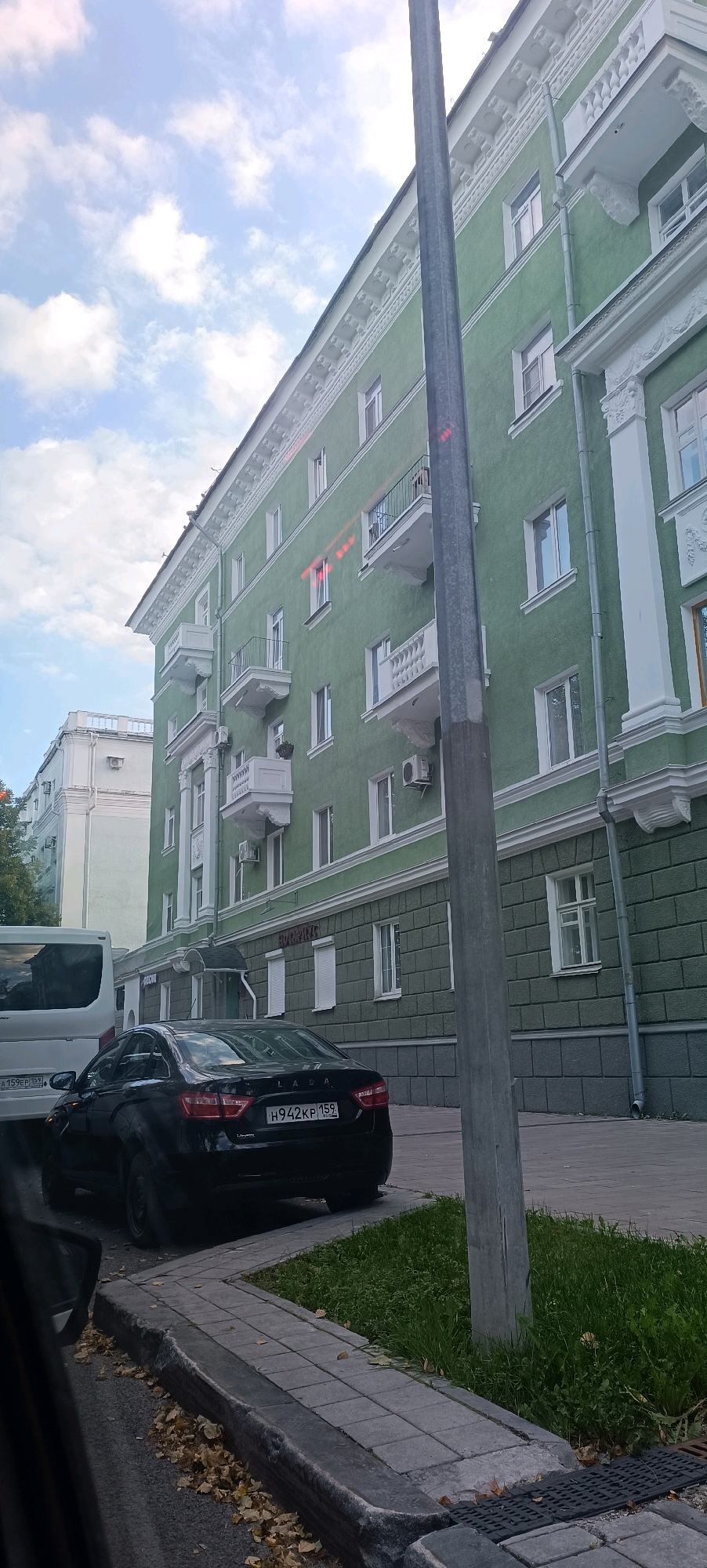 The image shows a street scene in Perm, Russia, on a cloudy day. A tall, green apartment building with white decorative trim and balconies dominates the right side of the frame. A grey utility pole stands in front of the building, partially obscuring it. In the foreground, a black Lada sedan is parked on the street. The car has a Russian license plate that reads "H942KP 159". To the left of the sedan, the rear of a white van is visible. The sidewalk is paved with grey tiles, and there is a patch of grass and fallen autumn leaves. The building on the left appears to be lighter in color and also has multiple floors and windows. Signs are visible on the green building, one appearing to be for "АПТЕКА" (Apteka, meaning Pharmacy) and another for "ОБЬЕКТ" (Obiekt, meaning Object or Facility). The sky is partially obscured by clouds.