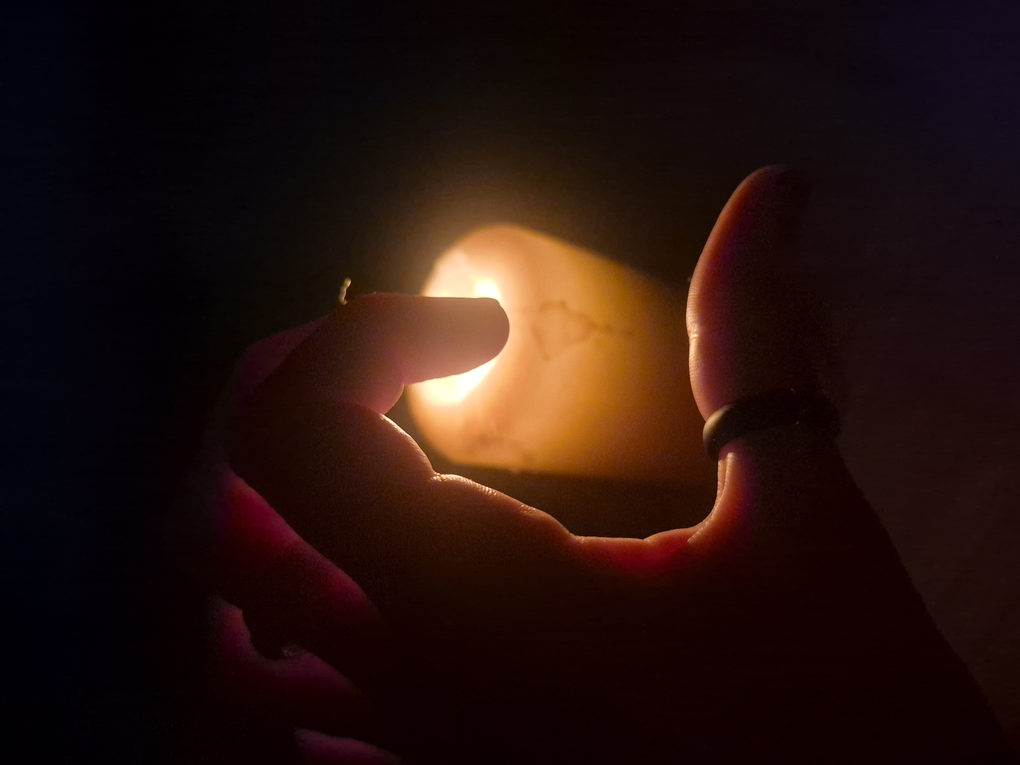 The image presents a close-up, low-angle shot of a human hand holding or positioned very near a lit candle in a dark setting, likely indoors or at night in Chaikovsky, Russia. The primary subjects are the hand and the candle, with the candle acting as the sole light source.
The hand is predominantly visible in profile, with fingers wrapping around the candle. An index finger and thumb are prominently featured on the left, close to the candle's glowing core. On the right side of the frame, parts of other fingers and the palm are visible, illuminated by the candle's warm light. A dark, simple band ring is worn on one of the fingers on the right side of the hand.
The activity depicted is the holding or interaction of a hand with a burning candle. The candle emits a strong, warm orange-yellow glow that powerfully illuminates the surrounding area immediately around it, creating bright highlights on the wax and backlighting the fingers closest to the flame. This light casts warm, reddish-orange tones onto the skin, creating a striking contrast with the deep black background. The flame itself is not directly visible, but its intense light source is. The overall atmosphere is one of intimacy and warmth, underscored by the strong chiaroscuro effect. There is no visible text in the image.