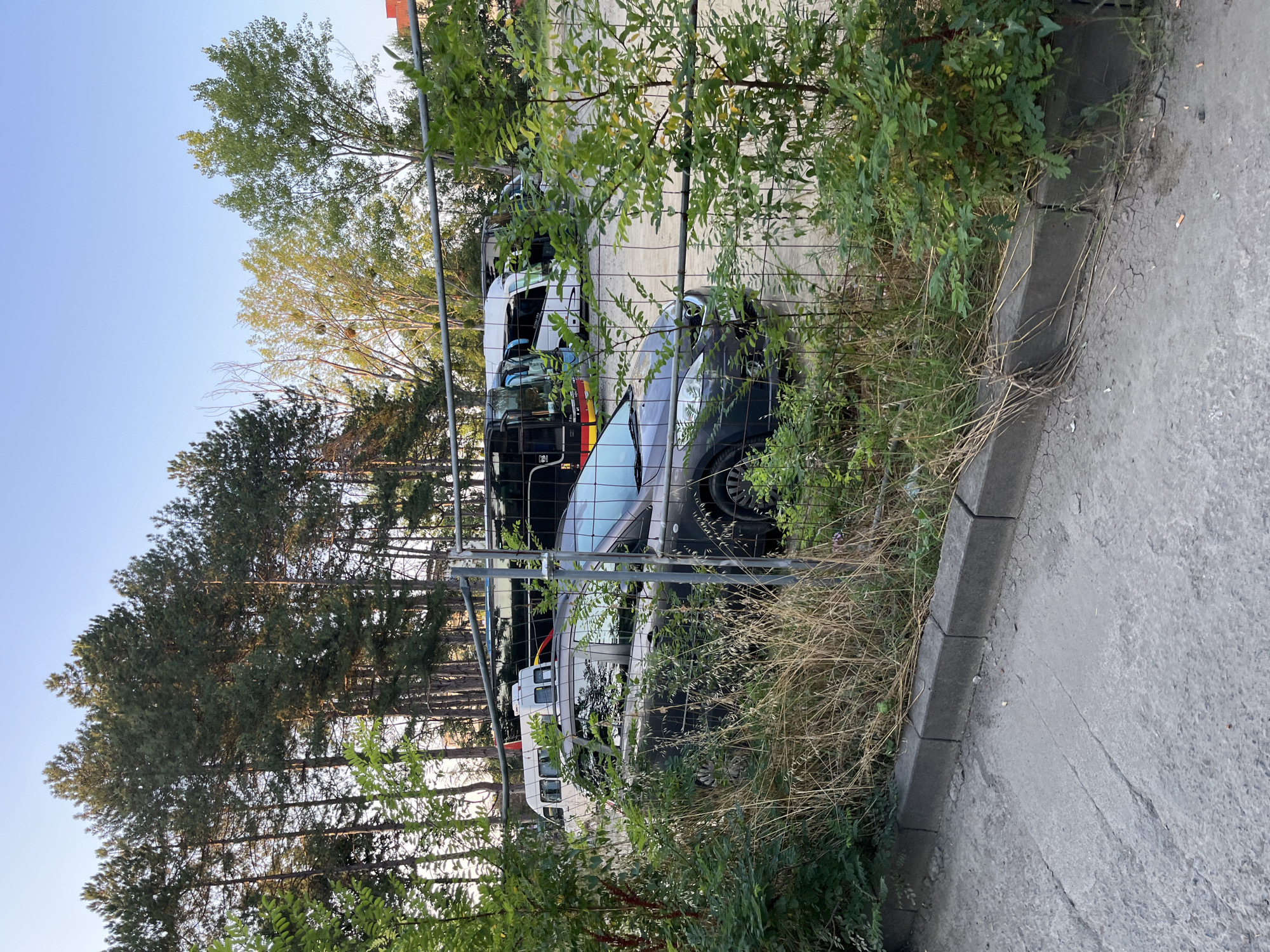 In Nesebar, Bulgaria, a dark-colored station wagon is parked behind a chain-link fence. The vehicle is partially obscured by tall grass and green foliage. Beyond the car, several buses are visible, parked in a lot. The sky above is clear and blue. The scene suggests a parking area, possibly for tour buses, located near a wooded area.