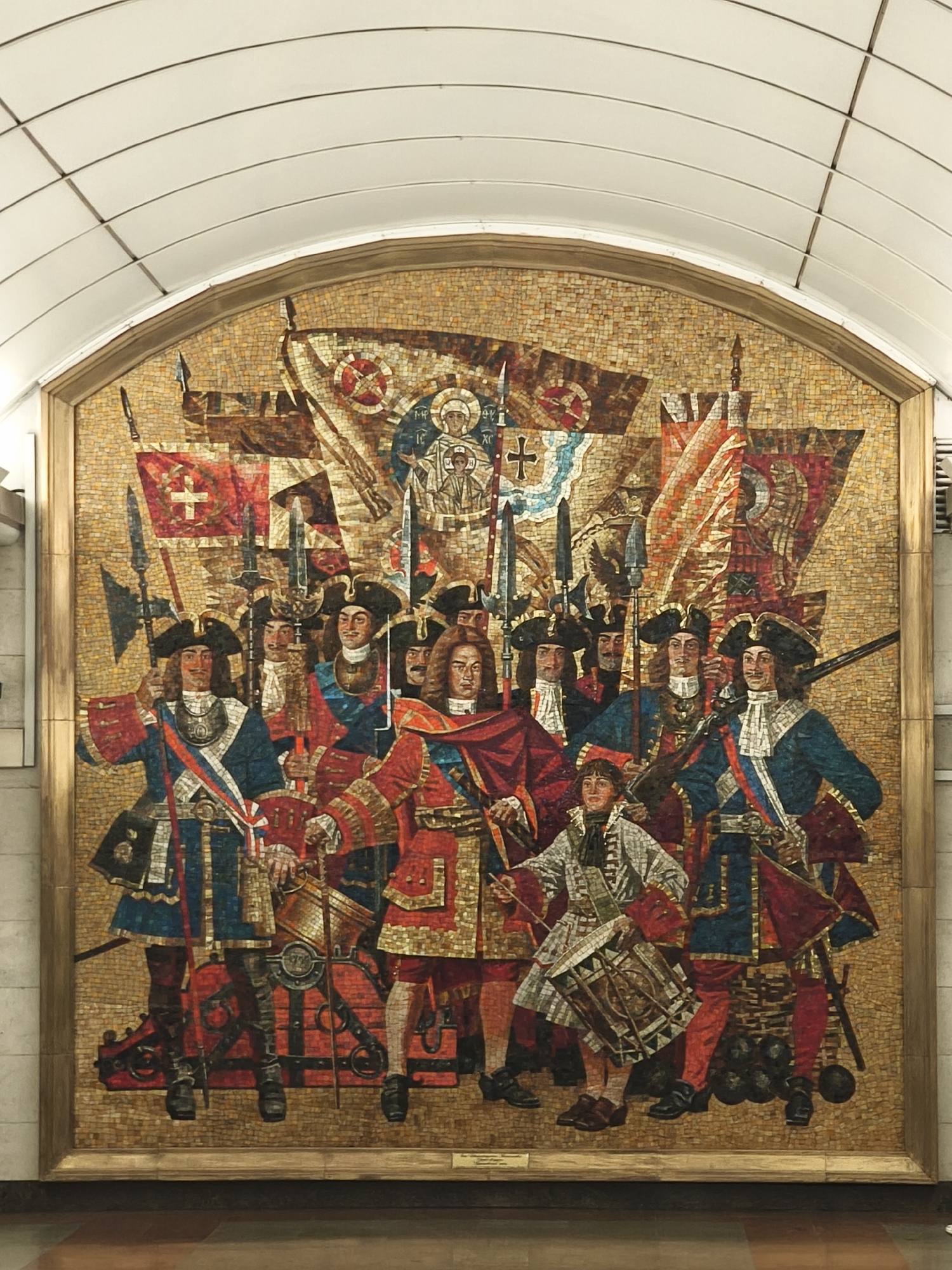 This is a mosaic artwork located in the Saint Petersburg Metro, likely within a station. The mosaic depicts a group of soldiers in historical military attire, possibly from the 17th or 18th century. They are dressed in elaborate uniforms with coats, breeches, stockings, and tricorn hats. Many of the figures are holding weapons such as spears and swords. In the center, a prominent figure in a red and gold coat appears to be a leader. A younger figure in the foreground is depicted playing a drum. Behind the soldiers, various flags and banners are displayed, some featuring religious iconography, including a depiction of Christ. The overall impression is of a military procession or a historical battle scene. The mosaic is framed by a golden border and set against a textured, golden background composed of small tesserae. The artwork is installed in an arched recess within a wall, suggesting it is a permanent fixture in a public space like a metro station.