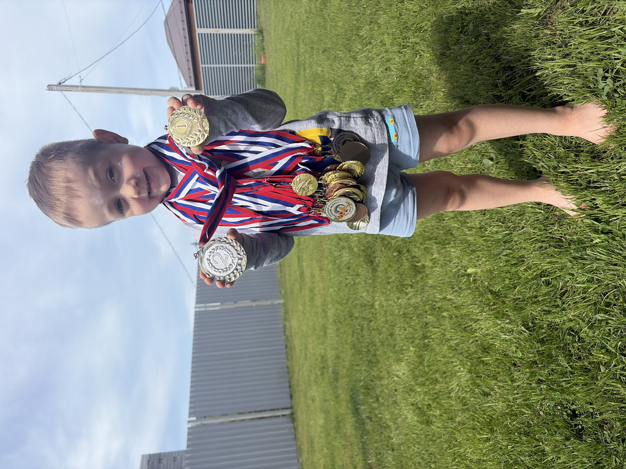 A young boy stands barefoot on a patch of green grass, holding a collection of medals. He is fair-skinned with short, light-colored hair and is wearing a gray long-sleeved shirt and blue shorts with a small car design on the side. The medals are various shades of gold, silver, and bronze, suspended from ribbons of varying colors, primarily red, white, and blue. He holds a gold medal in one hand and a silver medal in the other, displaying them towards the camera. In the background, there is a corrugated metal fence and a building with a brown roof. The sky is visible with a gradient of blue and light clouds. The location is possibly in the city of Chaikovsky, Russia.
