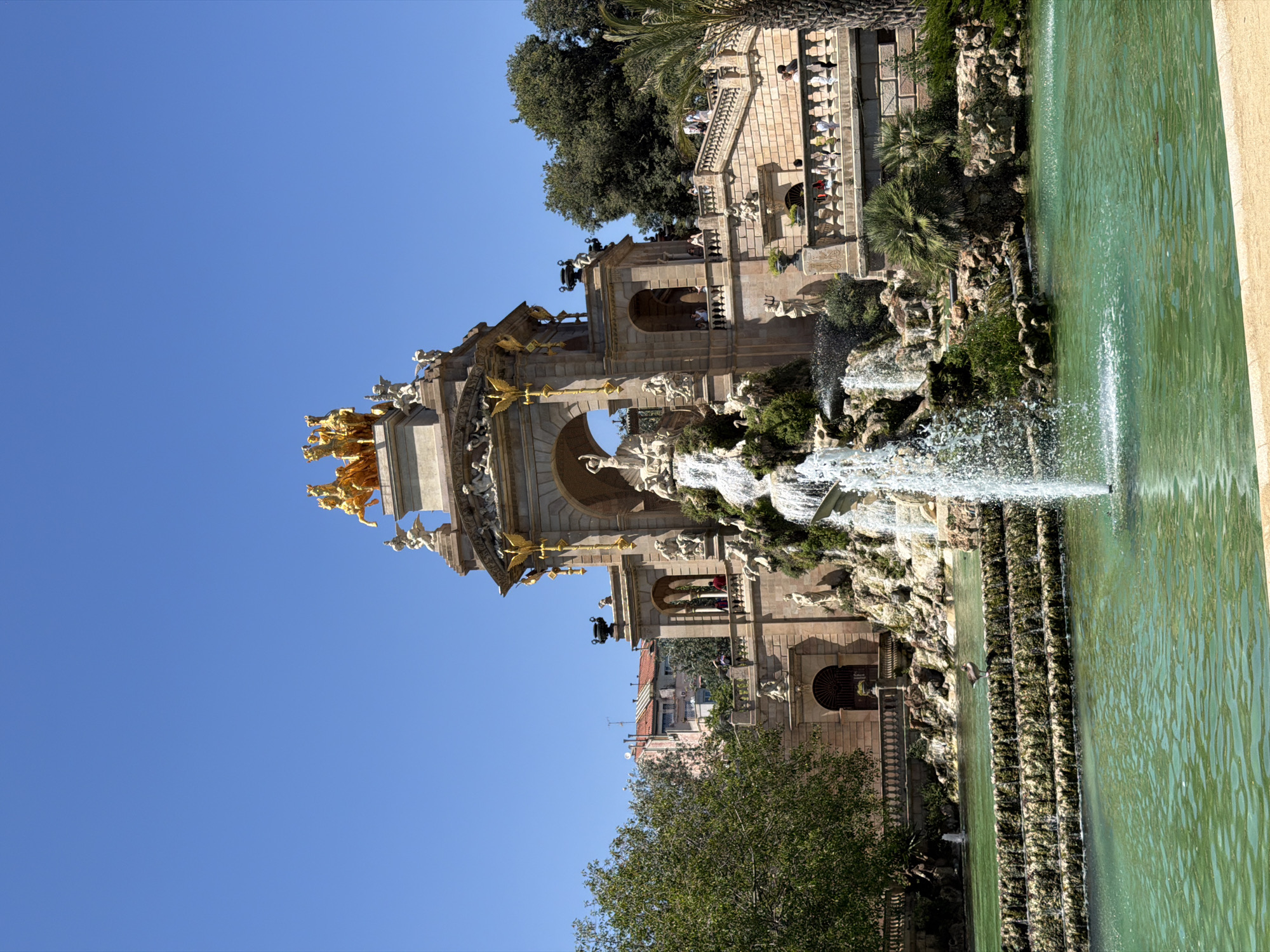 The image captures the Monumental Cascade in Parc de la Ciutadella, Barcelona, Spain. This elaborate fountain features a grand structure with golden sculptures of horses and a chariot at the top, set against a clear blue sky. Water cascades down a rocky, verdant structure, creating a dynamic and picturesque scene. People are visible on the balconies and stairs of the monument, observing the fountain. The foreground is dominated by a large, tranquil body of turquoise water, with a smaller fountain actively spraying water in the center. Lush green trees and palm trees frame the monument and the water feature, adding to the park's vibrant atmosphere. The architecture is ornate, with classical elements and sculpted figures adorning the stone facade. A lone bird stands on a tiered stone structure in the water.