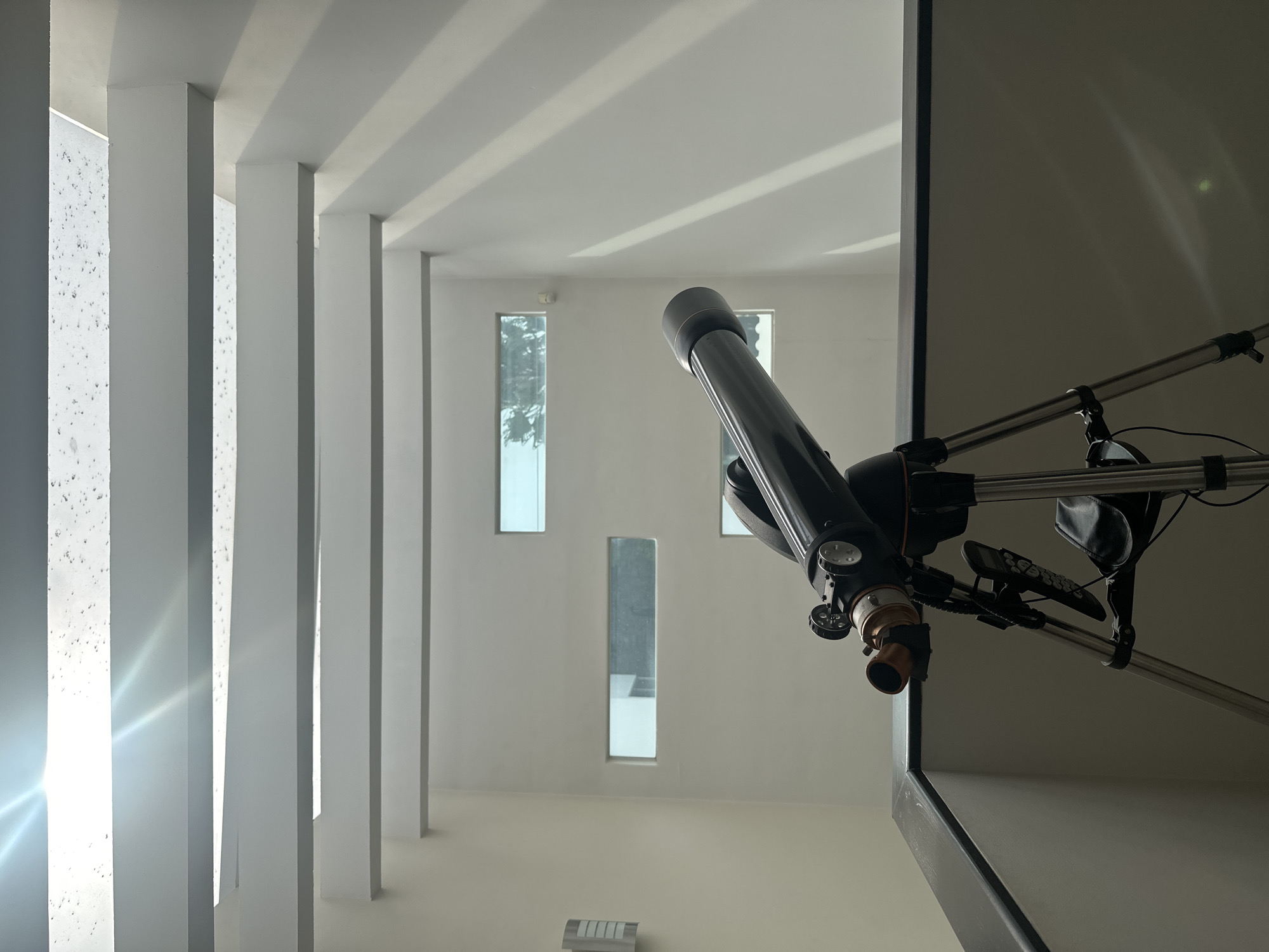A black telescope on a tripod stands on a dark railing, facing forward. The telescope has an orange accent on its eyepiece and a black tube. Behind the telescope, a multi-story indoor space with white walls is visible. Rectangular windows are set into the walls at different heights, and sunlight streams in from above through a slatted ceiling, casting diagonal shadows on the walls. The location is identified as San Bernardino Tlaxcalancingo, Mexico. There are no people present in the image.
