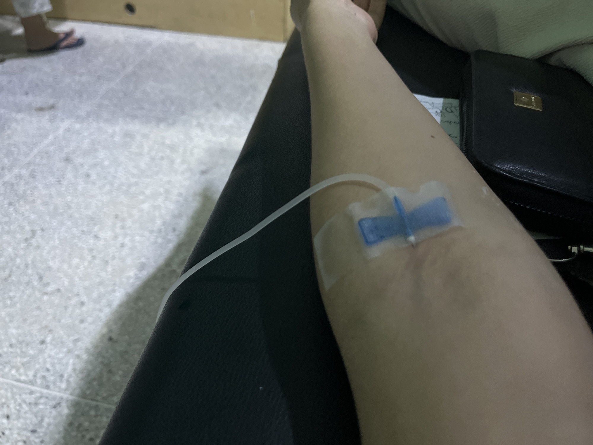 An intravenous catheter is inserted into the forearm of a person resting on a black leather-like surface. The catheter tubing is clear. A black wallet is next to the arm. The immediate environment appears to be indoors. The location of Juan Griego, Venezuela, cannot be determined from the image.