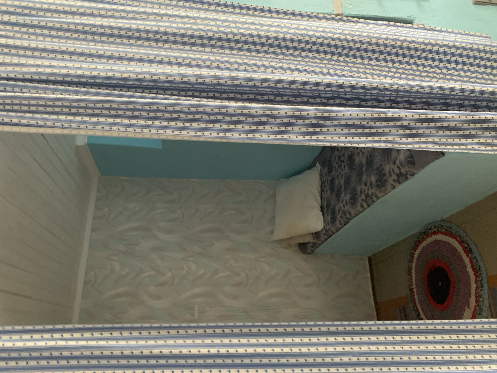 A narrow, bed-like structure with a light blue base and a patterned mattress is situated in a corner of a room. A cream-colored pillow rests on the mattress. The walls are covered in patterned wallpaper and a solid light blue paint. The ceiling is paneled with wood. A striped blue and white curtain hangs to the left and right of the bed, partially obscuring the view of the room. A circular rag rug with concentric rings of various colors is placed on the floor in front of the bed. The environment suggests a simple living space, possibly a bedroom or a nook. The location is identified as Відзаўскі сельскі Савет, Belarus. There are no people visible in the image.