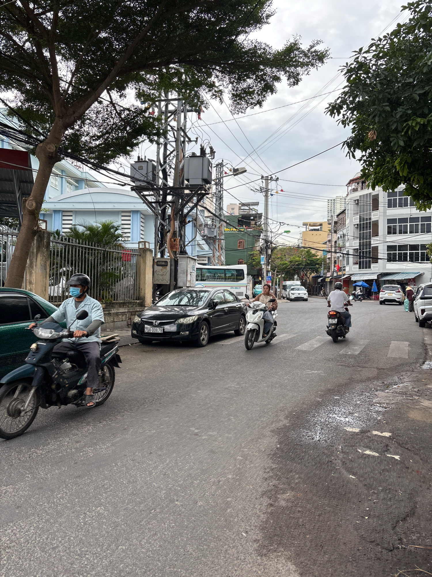 This is an eye-level, medium shot of a street scene in Nha Trang, Vietnam, on a cloudy day. In the foreground, a man wearing a light blue shirt, dark pants, a helmet, and a light blue face mask rides a dark colored motorcycle from left to right. The motorcycle is slightly angled towards the viewer. To the left of the motorcycle, a portion of a dark green car is visible. To the right of the motorcycle, a black sedan with license plate "79A-008.78" is parked.
The street is paved with asphalt and has visible road markings, including a pedestrian crossing. In the mid-ground, more vehicles are present, including a white van, several cars, and other motorcycles. The background features a variety of buildings, ranging from a light blue building with white trim to multi-story structures with modern facades. Overhead, a complex network of electrical wires and utility poles crisscrosses the sky. Trees with dense foliage frame the top of the image. The overall impression is a busy urban street with typical Vietnamese traffic and infrastructure.