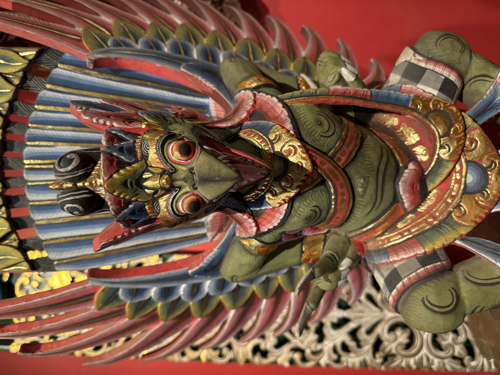 This is a close-up, overhead shot of a vibrantly colored, carved wooden sculpture of a mythical bird, likely a Garuda, a divine eagle-like creature in Hindu mythology. The sculpture is predominantly green, with accents of red, blue, gold, and black.
The bird's head is large and detailed, with piercing red eyes outlined in black and gold. Its beak is open, revealing a pink interior, and it has a golden crest. The bird is adorned with ornate gold detailing on its neck and chest, and it wears a traditional Balinese checkered cloth around its waist, a common motif in Indonesian art. Its wings are spread wide, composed of layers of carved and painted feathers in red, blue, and gold, giving a sense of powerful movement.
The sculpture appears to be part of a larger decorative element, possibly adorning a building or a shrine. The background is a solid red surface, likely a ceiling or wall, which makes the colors of the sculpture stand out. The lighting is bright, suggesting it might be daytime or under artificial lighting. The carving style is intricate and traditional, indicative of Indonesian craftsmanship, possibly from Bali, given the reference to Kuta, Indonesia as a location context. There is no visible text in the image. The overall impression is one of grandeur and religious significance, typical of Balinese art.
