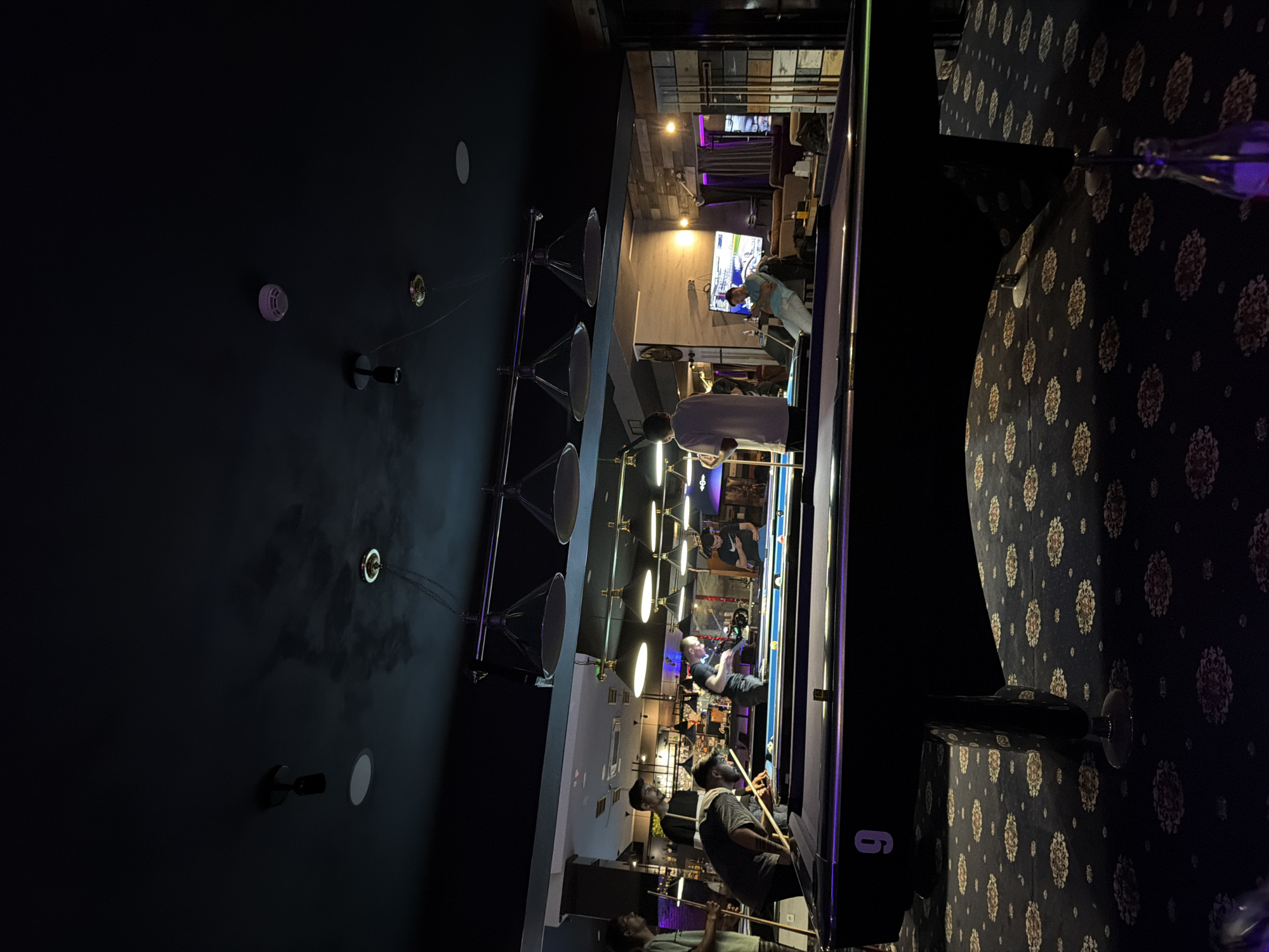 A photograph taken at night inside a billiards hall or pool hall, likely in Plovdiv, Bulgaria, shows a billiards table surrounded by people playing and watching. The table is lit by overhead lamps hanging from a rack system. Individuals are engaged in playing pool, holding cues, and aiming at billiard balls. The room has a modern, stylish interior with dark walls and patterned carpeting. Tables and seating areas are visible around the pool tables. Some people are standing while others are seated. A television in the background shows a sports game or program. The number "6" is visible on the side of one of the pool tables.