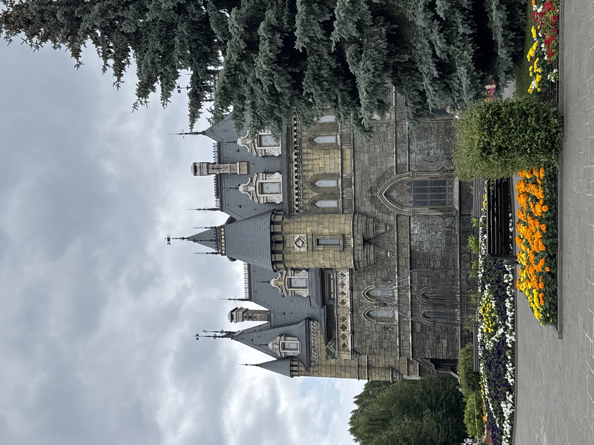 The image captures a view of the Khryashchevka Castle, located in Khryashchevka, Russia, under a cloudy sky. The castle, a prominent structure, is characterized by its gothic architectural style, featuring multiple towers, steep roofs, and stone masonry. A large evergreen tree stands to the right of the castle, partially obscuring its facade. In the foreground, a well-maintained flower bed displays vibrant orange, yellow, white, and purple blossoms, with a dark-colored park bench situated behind it. The ground is paved with interlocking grey stones. The overall scene depicts a picturesque and somewhat moody atmosphere, with the castle serving as the central subject against a backdrop of lush greenery and overcast skies.