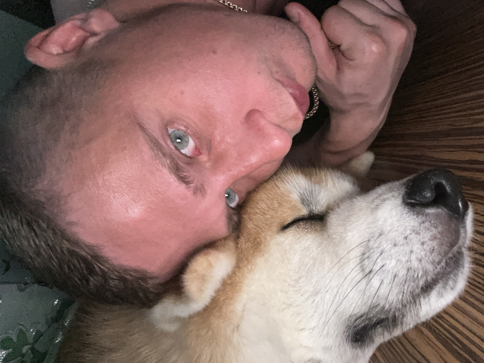 A close-up selfie shows a person and a dog lying down together. The person, with short, dark hair and light blue eyes, rests their head on the dog's body. The dog, a Shiba Inu with tan and white fur, has its eyes closed and is nuzzling into the person's cheek. They are lying on a wooden surface, possibly a floor or table. The background is dark and out of focus, with some hints of green foliage. There is no discernible location in Minsk, Belarus.