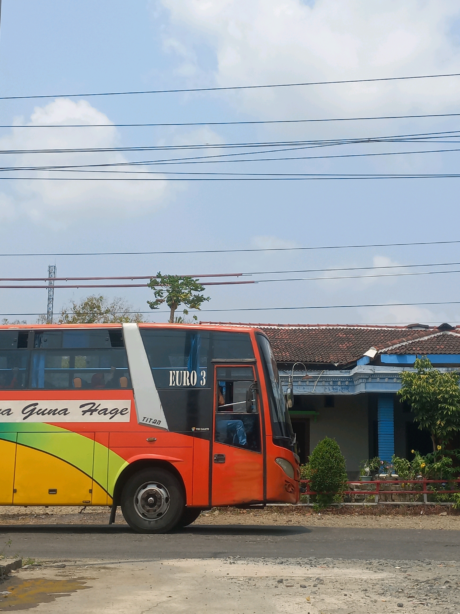 An orange bus with colorful stripes on its side drives on a road in Baturetno, Indonesia. The bus has the text "EURO 3" printed on its side. A building with a tiled roof and blue trim is visible behind the bus, along with trees and vegetation. Several power lines stretch across the sky. The ground is paved with asphalt and gravel. The sky is blue with some clouds.