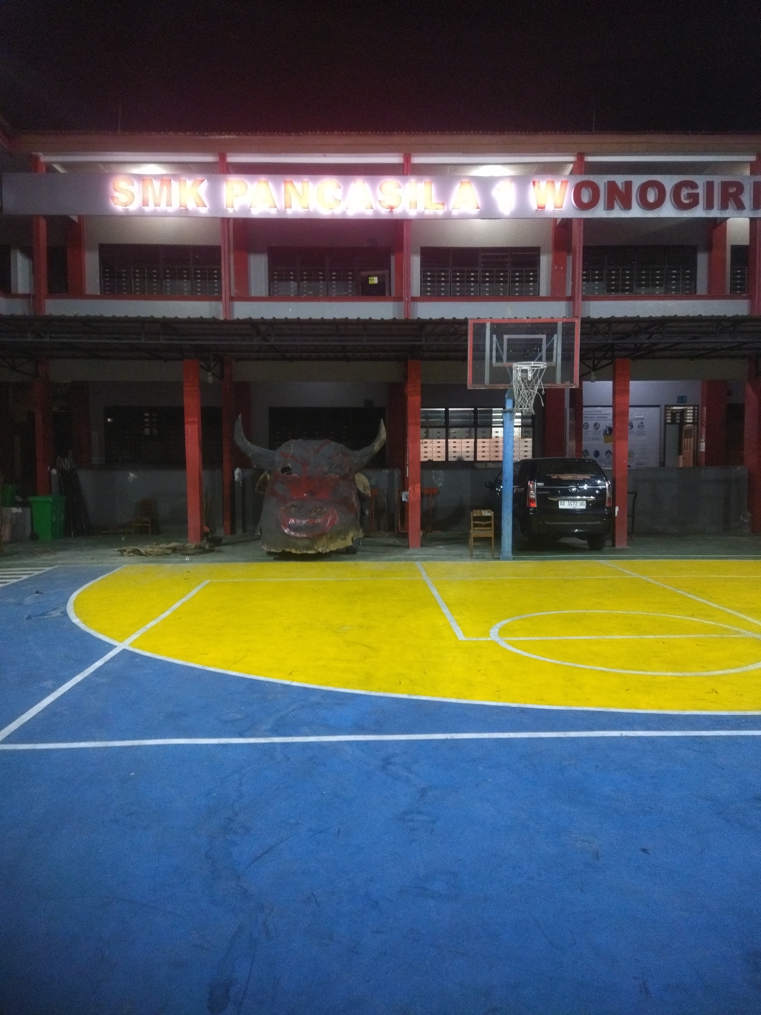 The image depicts an outdoor basketball court with a blue and yellow surface. In the background, a two-story building with red accents and windows is visible. A large, dark-colored effigy of a bull's head with red markings and horns stands on the court near the building. A basketball hoop is positioned on a pole. Behind the bull effigy, a black SUV is parked, and a small wooden chair is placed near it. The scene appears to be illuminated by artificial lighting, suggesting it is nighttime. The building has a prominent sign in illuminated letters that reads "SMK PANCASILA WONOGIRI." The location is identified as Giritirto, Indonesia.