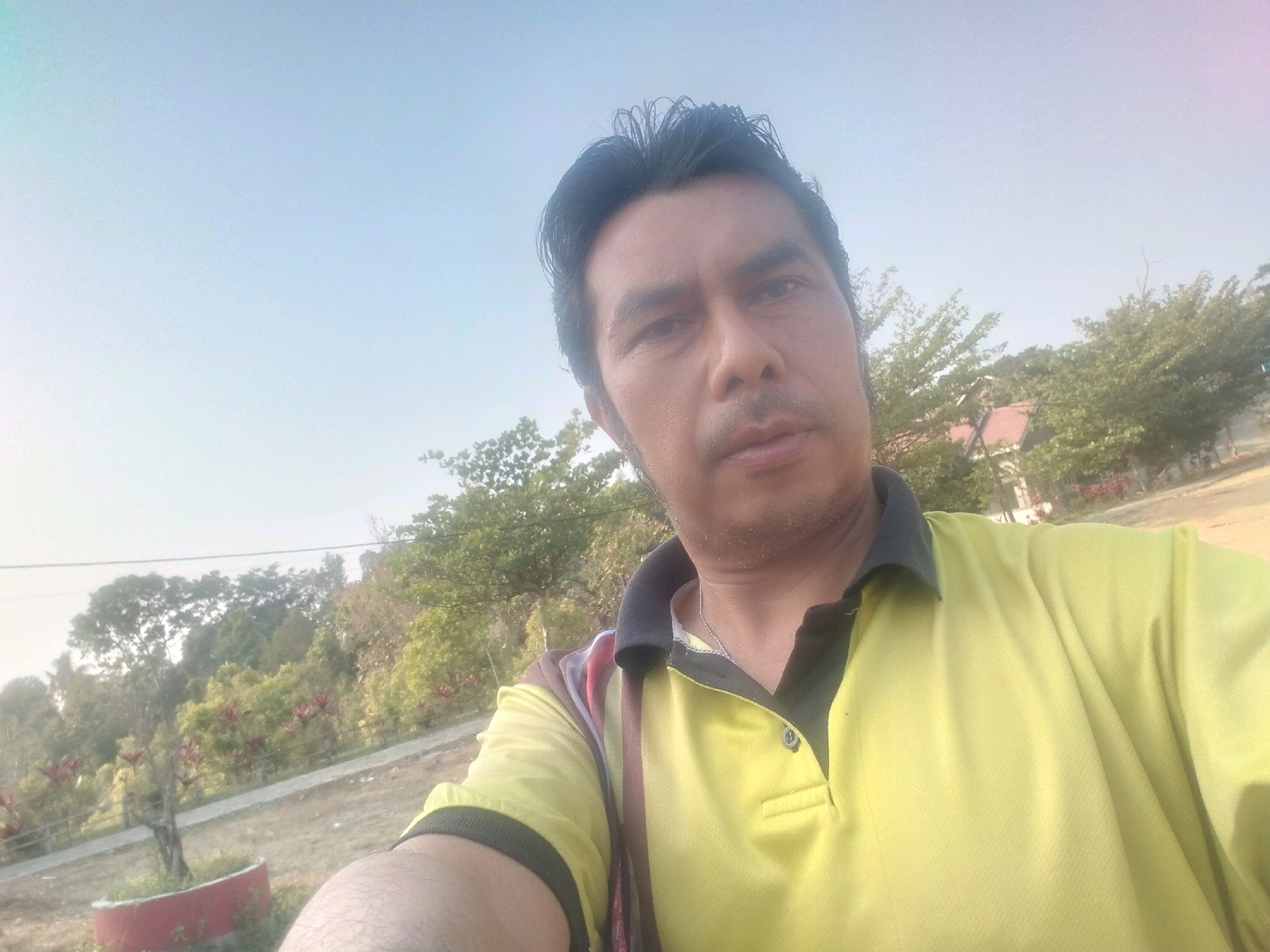 A man takes a selfie outdoors in what appears to be a park-like setting. He is wearing a bright yellow polo shirt with a black collar and trim. The background shows trees, foliage, and a clear sky. The image is taken from chest-up, with the man's face prominently featured. He has dark hair and is looking directly at the camera. The lighting suggests it is daytime. The location is identified as Sawahan, Indonesia.