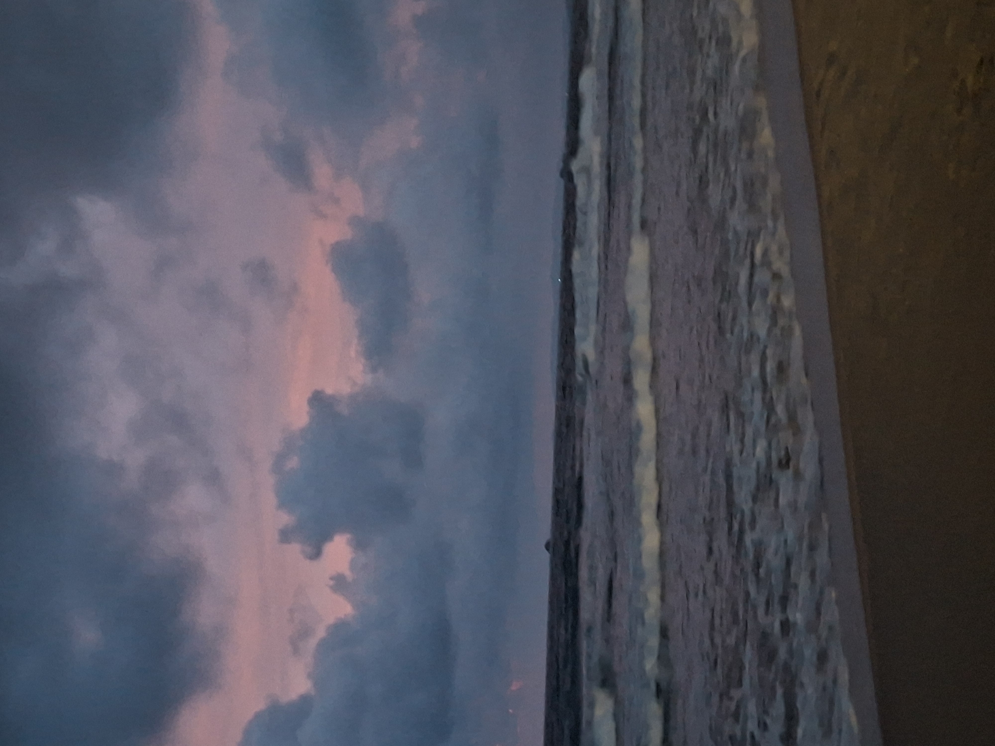 The image depicts a tranquil beach scene at dusk on Phú Quốc, Vietnam. The sky is painted with hues of soft pink and deep gray clouds, creating a dramatic yet serene atmosphere. The sea gently laps against the shore, with waves creating white foam as they break. In the distance, faint outlines of land or islands are visible on the horizon. The foreground is occupied by a dark sandy beach. No people or specific landmarks are identifiable, contributing to the overall sense of natural beauty and isolation.