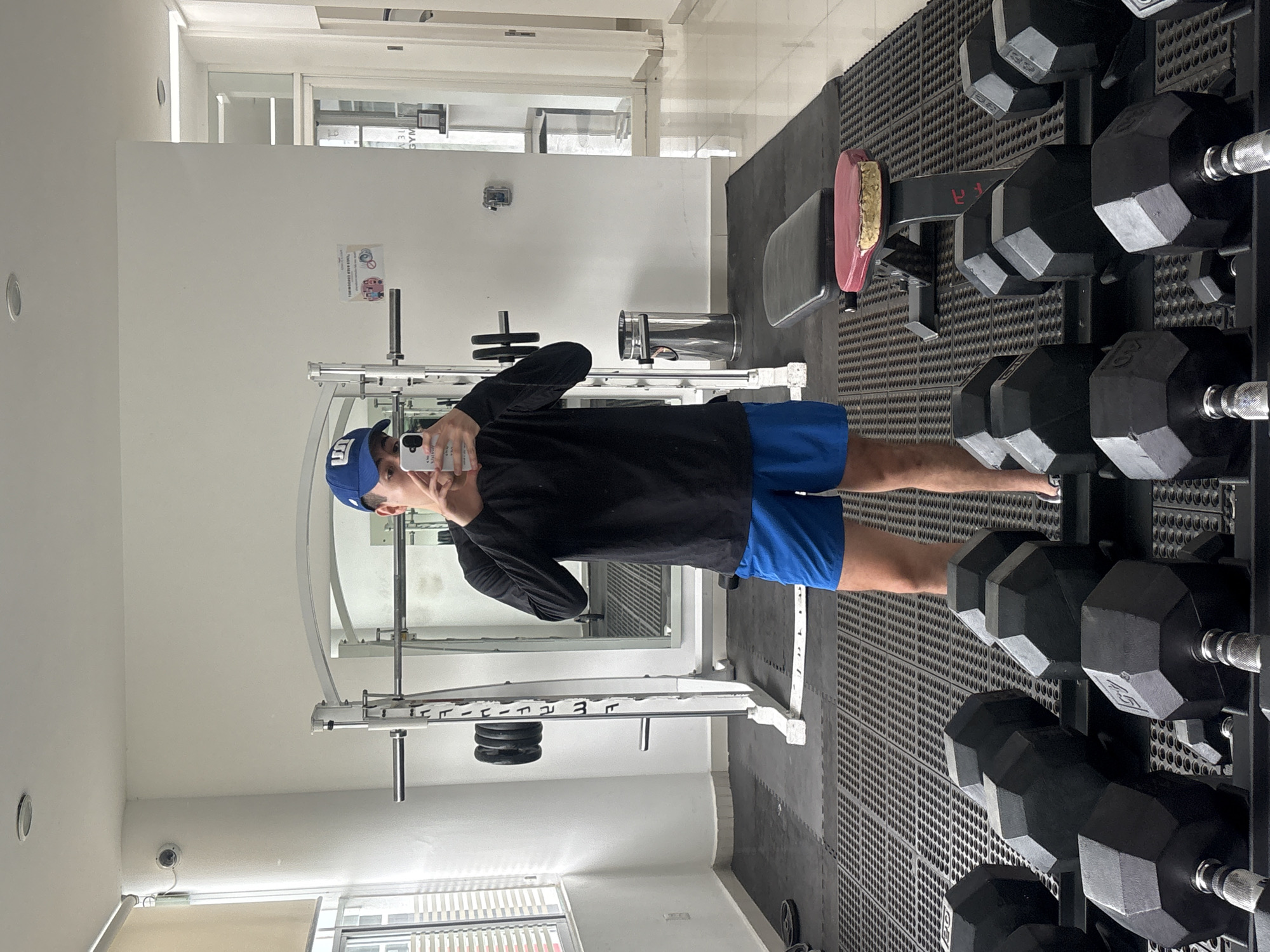 A young man takes a mirror selfie inside what appears to be a gym during the daytime. He is standing barefoot, wearing a black long-sleeved shirt, blue shorts, and a blue baseball cap with a white logo (possibly "NY"). His phone obscures part of his face as he holds it up to capture his reflection.
The gym environment includes various exercise equipment. To the right of the man, a black metal dumbbell rack holds several hexagonal dumbbells of different weights, with "40 LB" visible on one. A black and red/pink weight bench is positioned on a textured black rubber mat floor. Behind the man, a white squat rack or power cage with some black weight plates on its pegs is visible. A metallic trash can stands near the back wall.
The gym has white walls and bright lighting, suggesting either strong artificial lights or natural light from windows, which are visible in the background, reflecting the word "GYM." A small security camera is mounted high on the left wall. A sign on the wall contains pictograms and partially visible text, including "THANK YOU FOR YOUR COOPERATION." Other text like "GYM" is visible reversed in reflections.