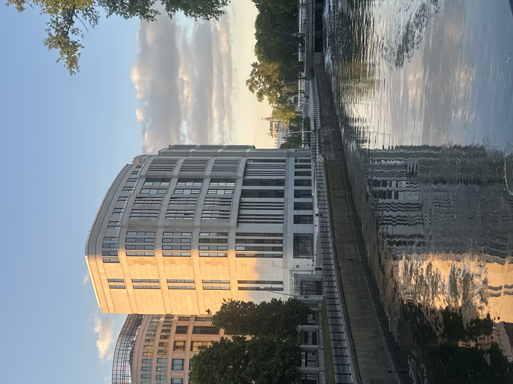 The image captures an urban waterside scene, likely in Minsk, Belarus, during the golden hour. A large, modern building with a curved façade dominates the left side of the frame, its light-colored stone or concrete exterior warmly illuminated by the setting or rising sun. This building features numerous rectangular windows arranged in horizontal bands. A similar, partially visible building stands behind it.
To the right, a calm body of water, presumably a river or canal, stretches into the distance, reflecting the sky, clouds, and structures on its banks. Gentle ripples are visible on the water's surface. Along the far bank of the waterway, a paved walkway or embankment is discernible, lined with trees and sparse vegetation. In the background, beyond the trees, other urban structures are visible, including one with a distinct dome-like roof.
The sky is a clear blue, adorned with scattered cumulus clouds that are also touched by the warm sunlight. Trees with green foliage are visible in the upper left foreground, framing a portion of the sky, and also line the distant riverbank. The overall lighting suggests late afternoon or early evening, creating long shadows and a soft, warm ambiance. No people, specific activities, or discernible text are present in the image.