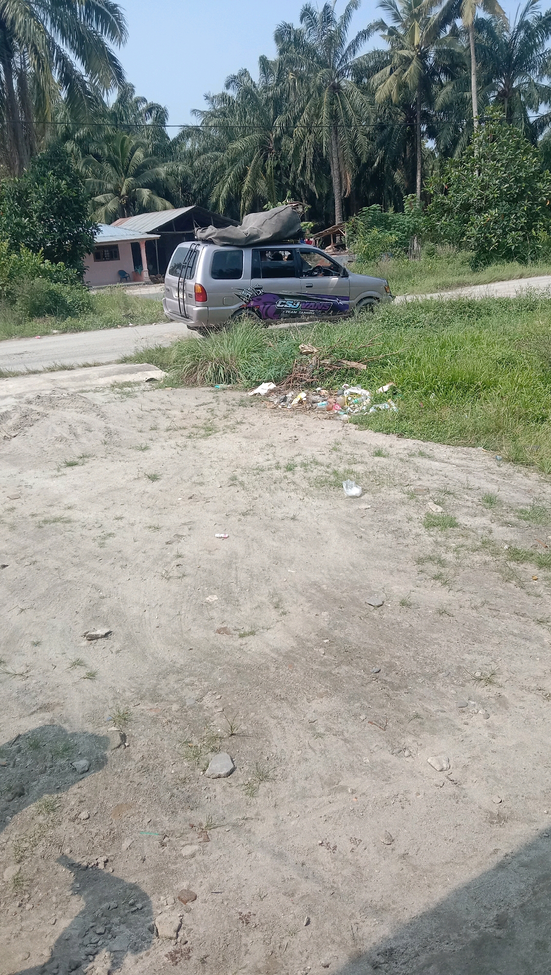 In Kota Pari, Indonesia, a silver minivan is parked on a dirt road beside a grassy area. The minivan has a large grey tarp strapped to its roof and a purple graphic sticker on its side that reads "CSU FAILS TEAM TAHUN". In the background, palm trees and a few buildings are visible. A pile of trash is located next to the minivan on the right side of the road. The ground in the foreground is a mixture of dirt and small rocks with sparse patches of dry grass. The sky is clear and bright.