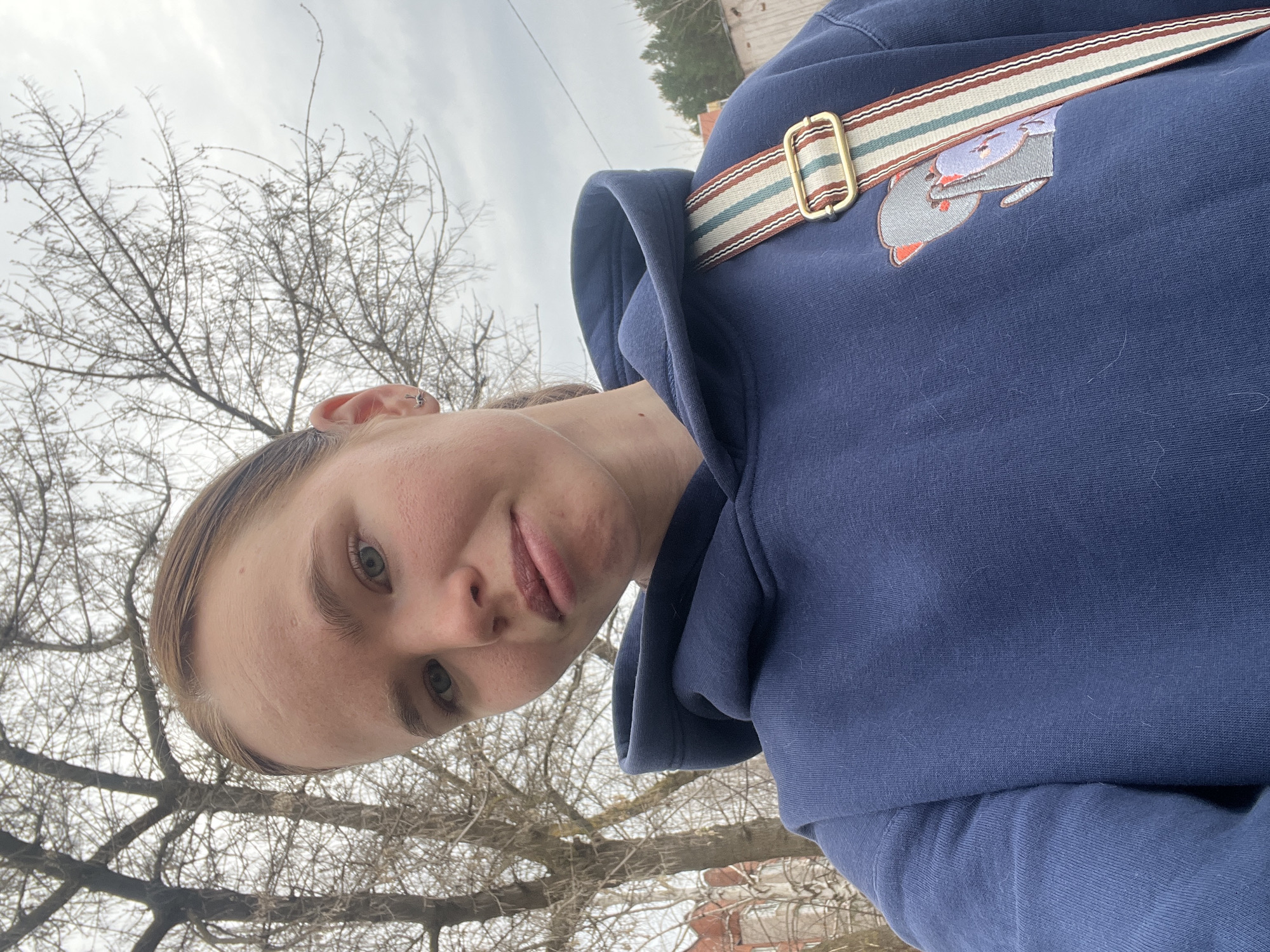 A woman with light skin and short blonde hair is taking a selfie outdoors. She is wearing a navy blue hooded sweatshirt with a small embroidered animal design on the upper right chest and a patterned strap with a gold-colored buckle slung across her chest. The background consists of bare tree branches against a cloudy sky. The location is Chaikovsky, Russia.