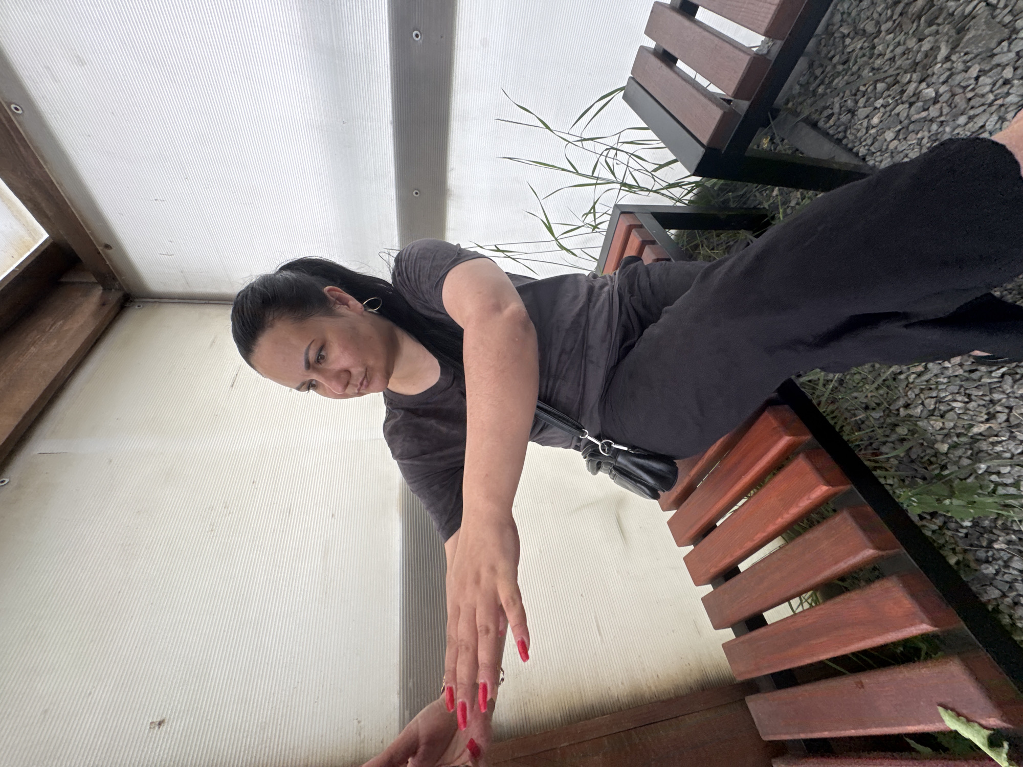 A woman with dark hair pulled into a ponytail sits on a wooden bench in what appears to be an outdoor covered area, possibly a greenhouse or a sunroom. The walls of the structure are made of corrugated translucent material, and wooden beams support the ceiling. The woman is wearing a dark grey t-shirt and black pants. She has long, bright red acrylic nails and wears a simple hoop earring in her left ear. A small black crossbody bag is slung over her right shoulder. Her left arm is extended forward, with her hand open, as if reaching out or presenting something. Another bench, similar in design with wooden slats, is visible to her right, and the ground is covered in small grey stones and some sparse green vegetation. The lighting suggests it is daytime. The location is identified as Zaliznychne, Ukraine.